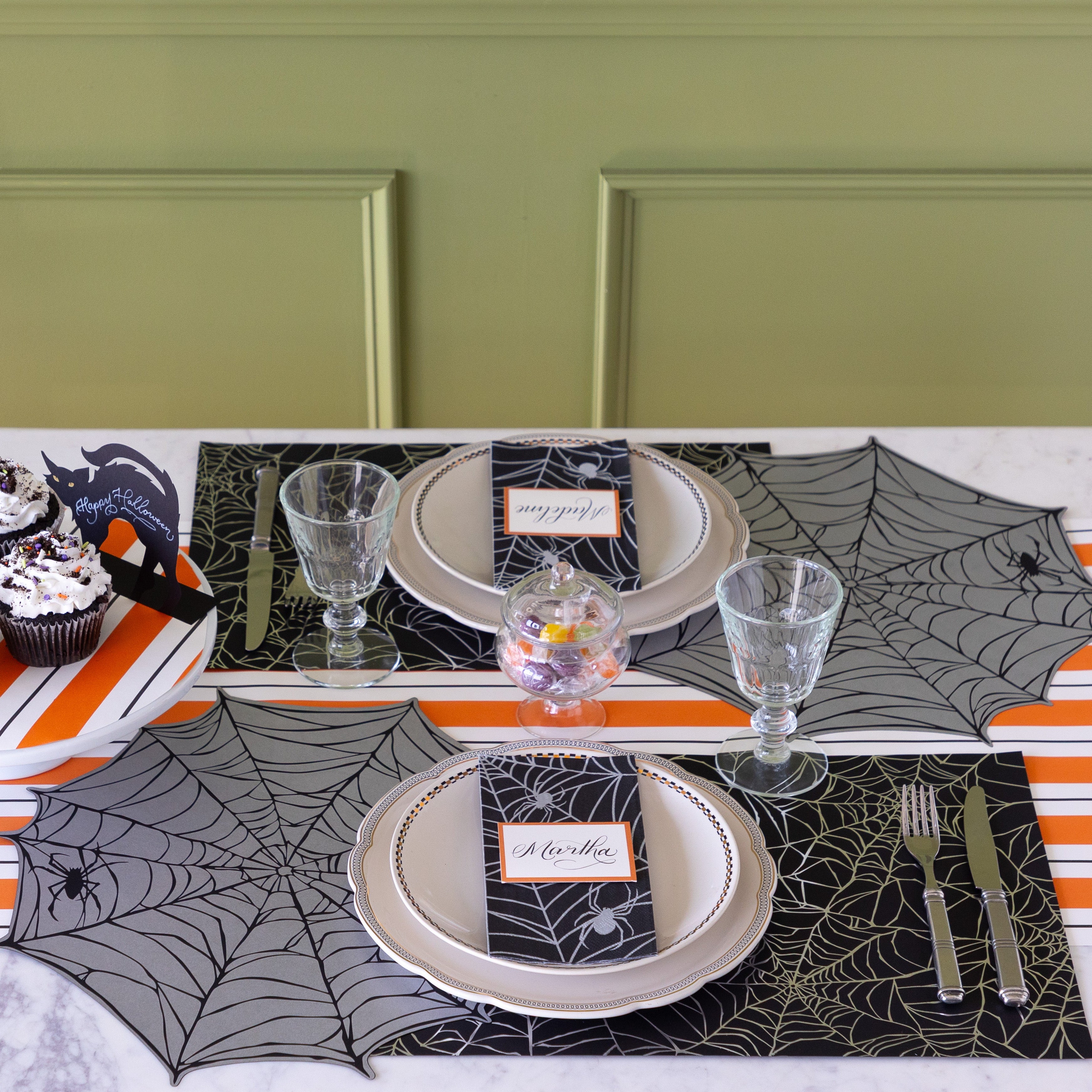 A Halloween themed table setting featuring the Die-cut Spiderweb Placemat slightly overlapping the Spiderweb Placemat, the Spiderweb Guest Napkin with Orange Frame Place Card on top and Orange &amp; Black Awning Stripe Runner underneath.