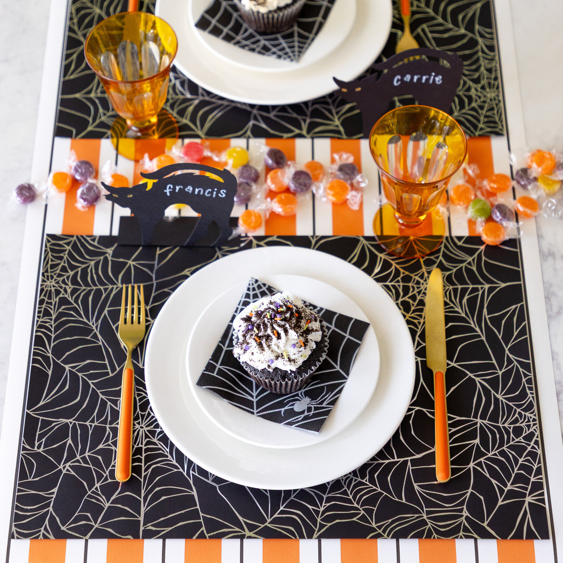 A Halloween table setting with candy featuring the Spiderweb Placemat with an orange stemmed, gold knife and fork, a cupcake on the Spiderweb Cocktail Napkin resting on the plates, Black Cat Place Card reading "Francis" and Orange &amp; Black Awning Stripe Runner underneath.