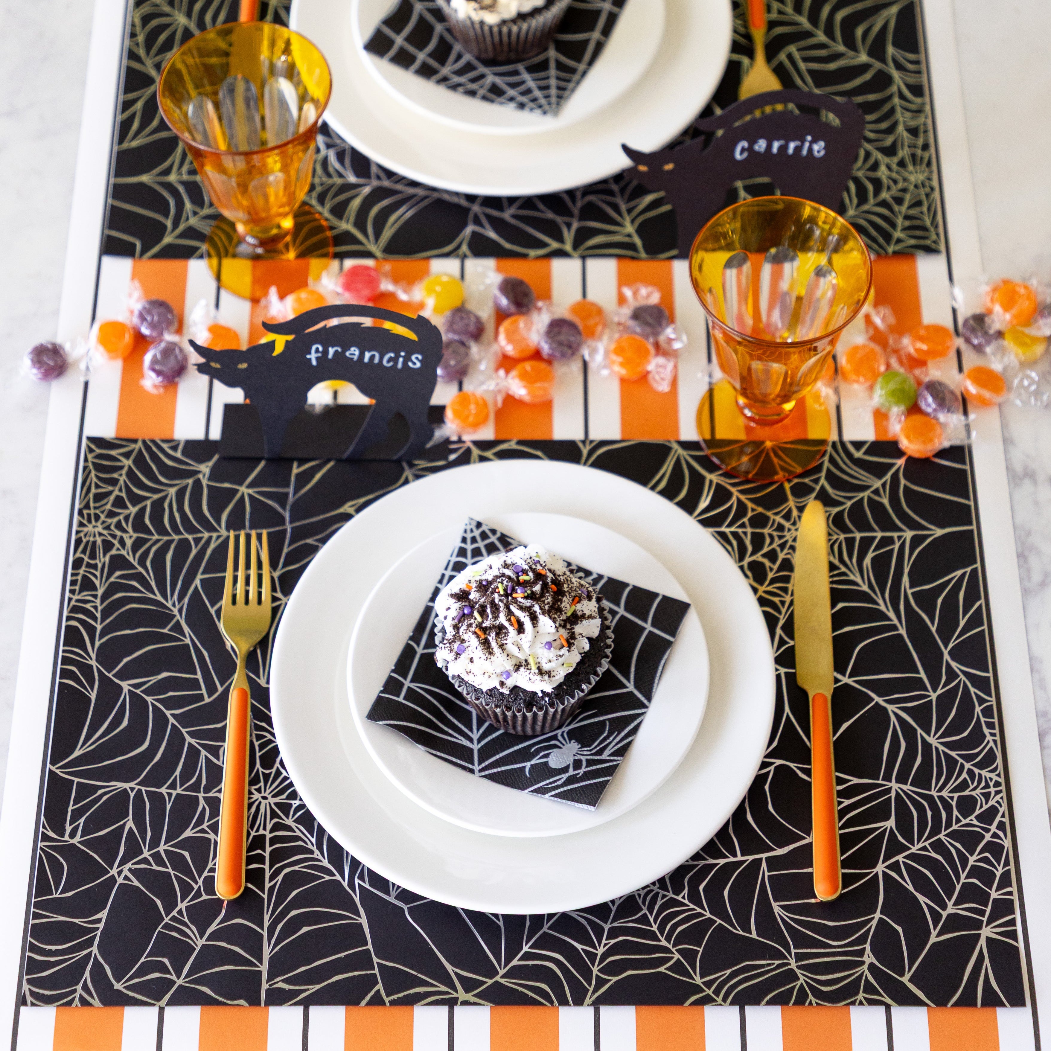 A Halloween table setting with candy featuring the Spiderweb Placemat with an orange stemmed, gold knife and fork, a cupcake on the Spiderweb Cocktail Napkin resting on the plates, Black Cat Place Card reading "Francis" and Orange &amp; Black Awning Stripe Runner underneath.