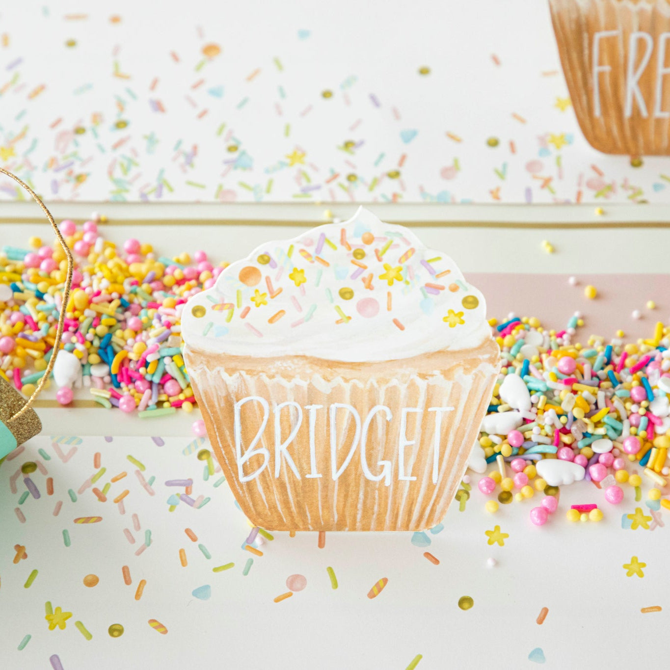 Cupcake Place Card with &quot;Bridget&quot; written on the wrapper part, on a table surrounded by sprinkles.