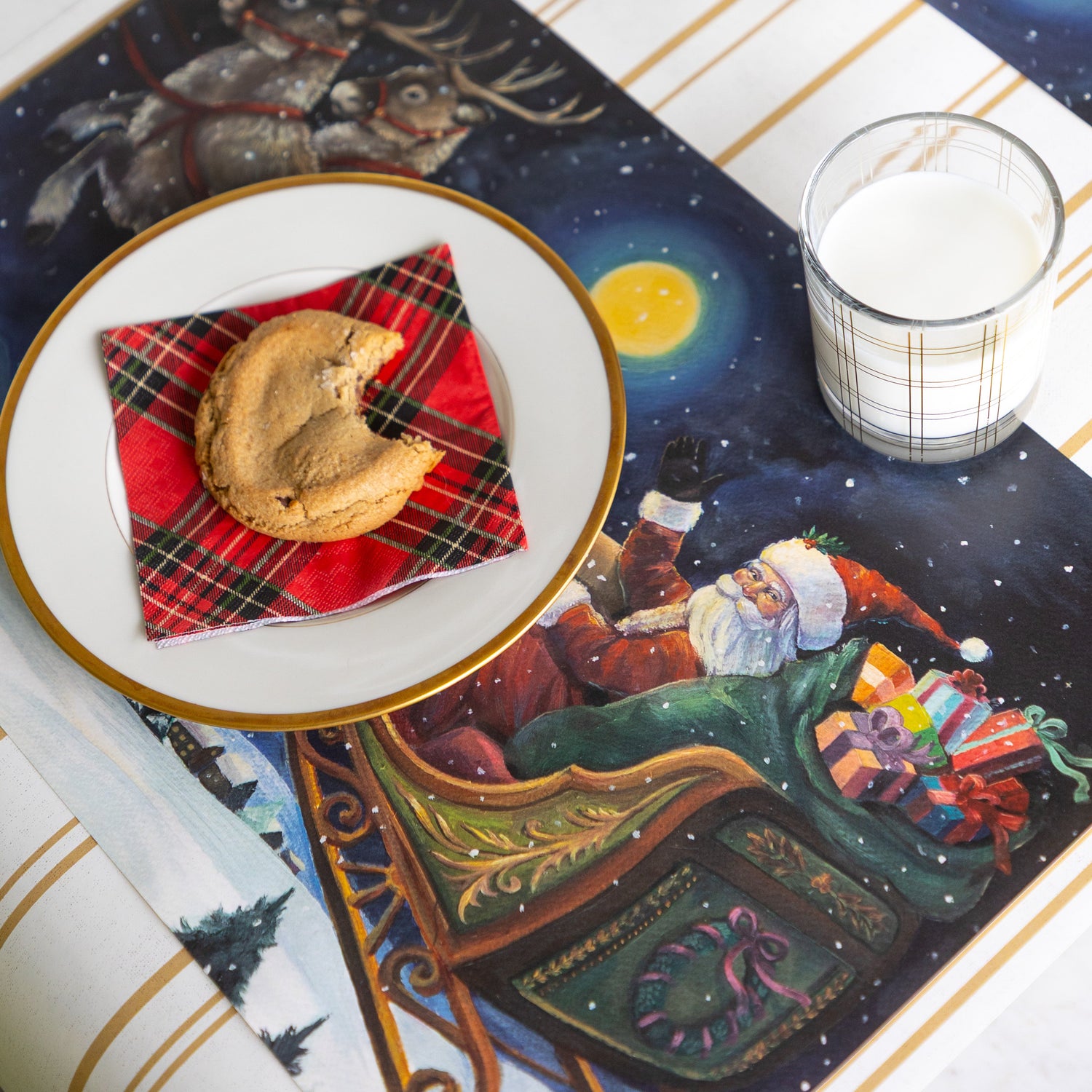 A place setting featuring the Sleigh Ride Placemat with a gold rimmed plate, Red Plaid Cocktail Napkin and cookie atop, a glass of milk and Antique Gold Stripe Runner underneath.