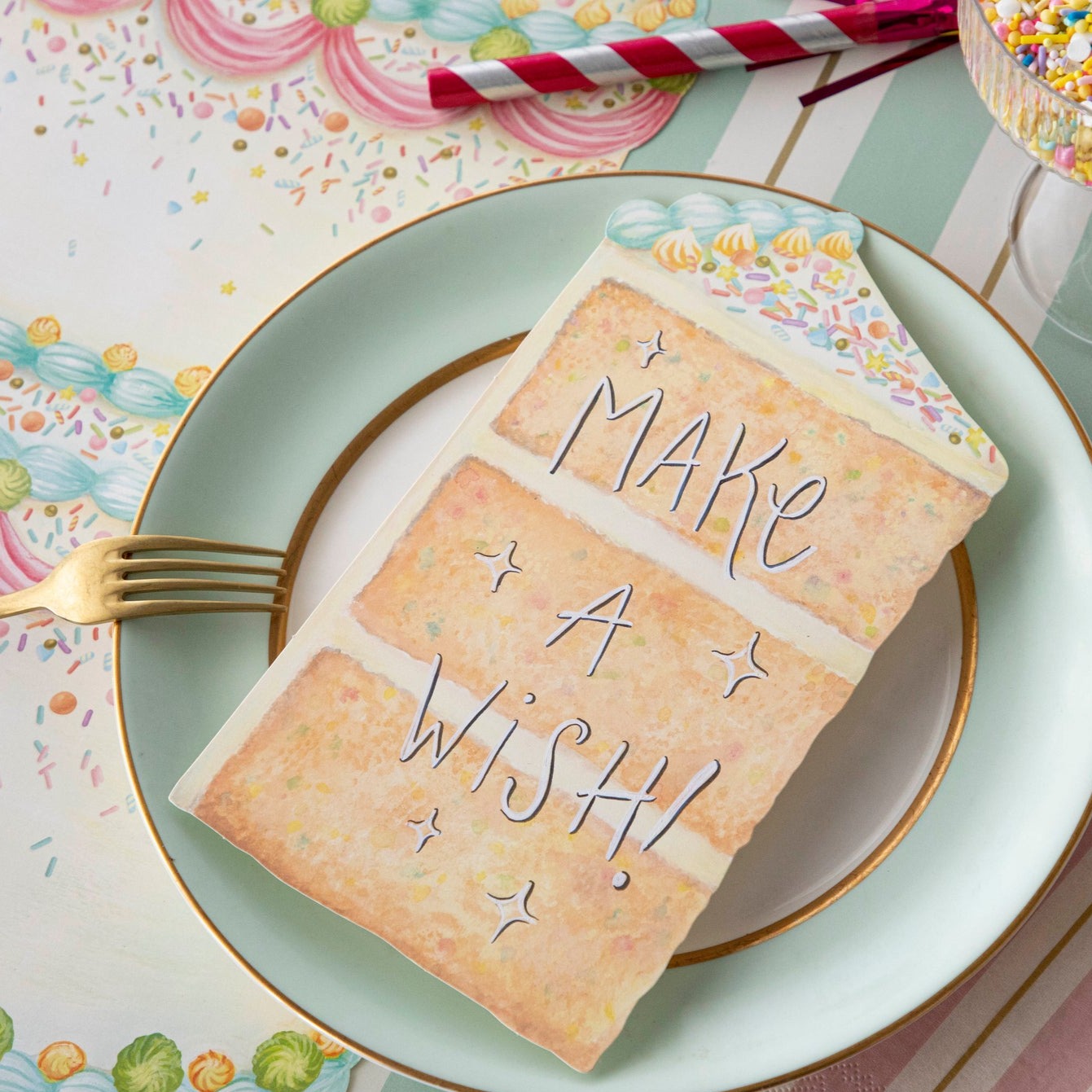 Close-up of a vibrant birthday party place setting featuring a Cake Slice Table Accent resting on the plate with &quot;Make A Wish!&quot; written in white ink with a black outline.