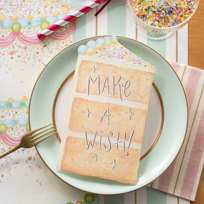 A vibrant birthday party place setting featuring a Cake Slice Table Accent resting on the plate with &quot;Make A Wish!&quot; written in white ink with a black outline, from above.