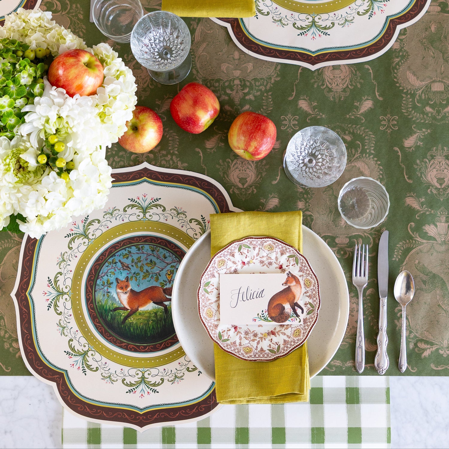 A decorative table setting with scattered apples and flowers, featuring the Die-cut Fanciful Fox China Placemat, vintage dinnerware with the Fanciful Fox Place Card on top and Moss Fable Toile Runner running horizontally and Dark Green Painted Check Runner running vertically on the table.