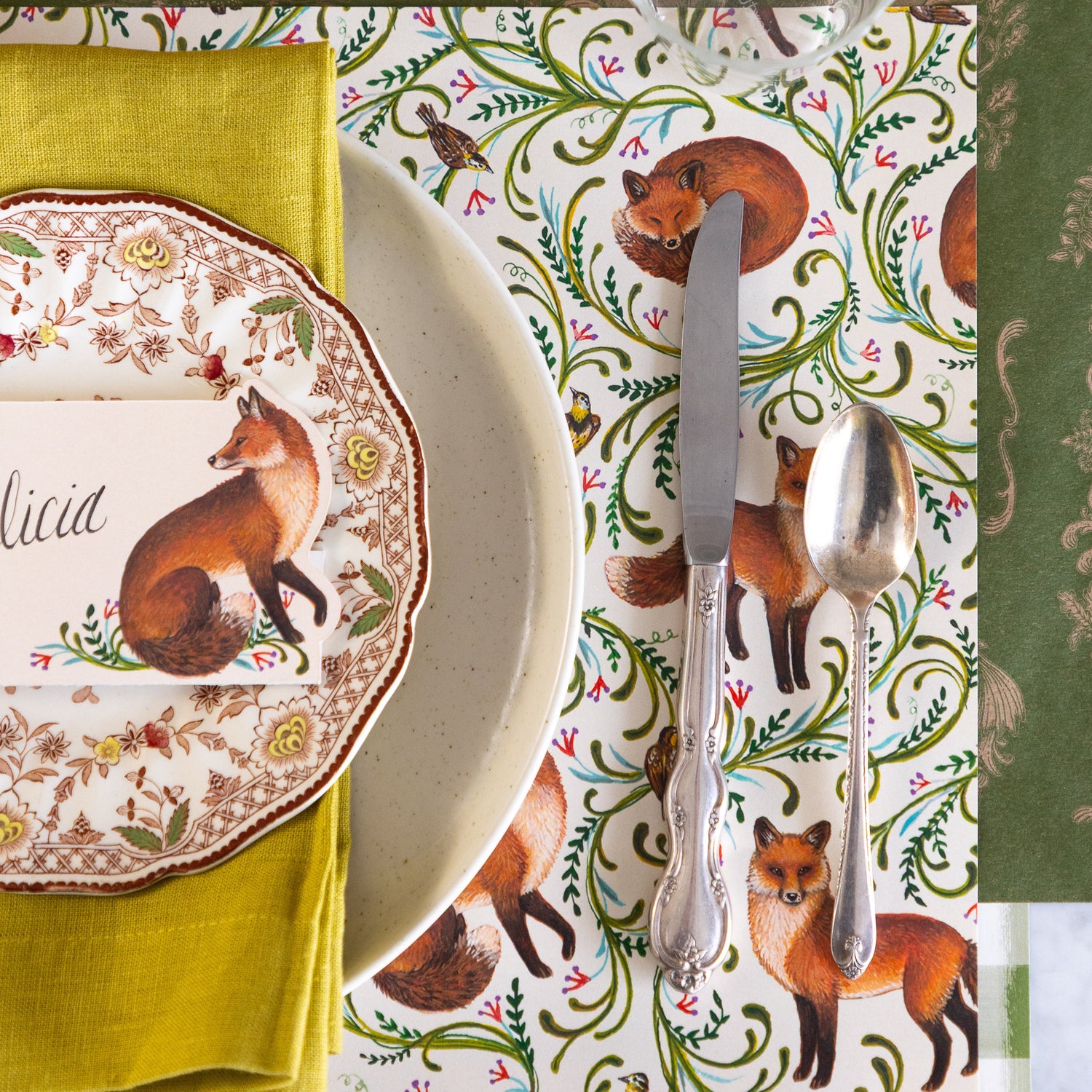 An elegant place setting featuring the Fanciful Foxes Placemat, vintage silverware, Pacifica Salt Dinner Plate with a green napkin, vintage floral plate and Fanciful Fox Place card on top, and Moss Fable Toile Runner under the place setting.