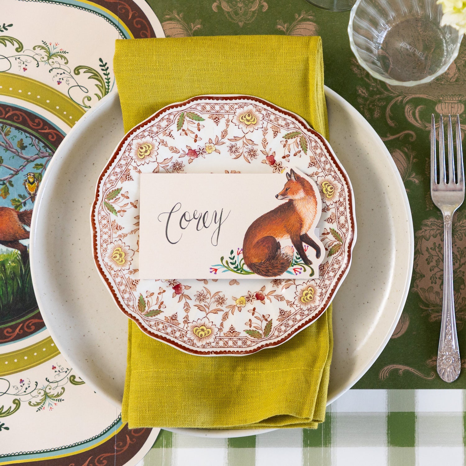 An elegant place setting featuring the Die-cut Fanciful Fox Placemat under a green napkin in between the Pacifica Salt Dinner plate and vintage floral salad plate with the Fanciful Fox Place Card reading "Corey" on top.