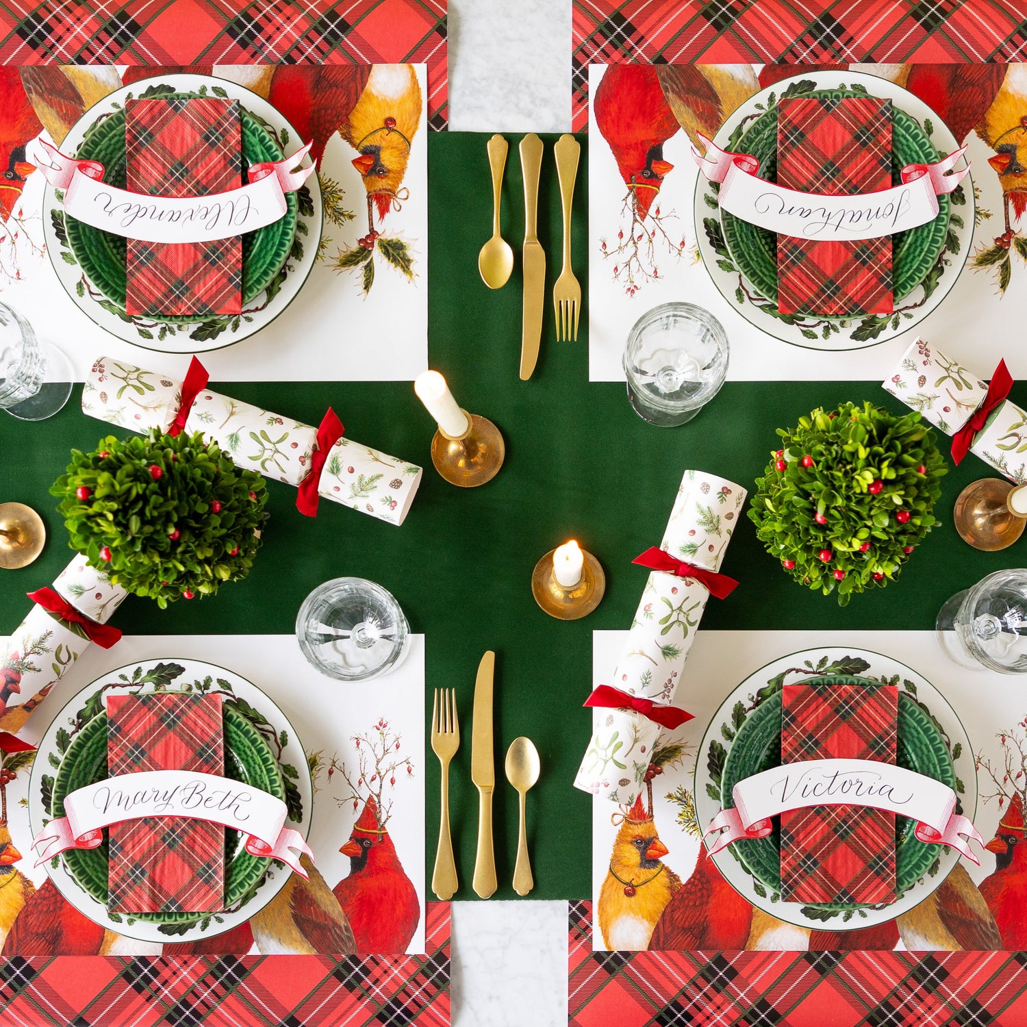 A festive table setting featuring the Green Flocked Runner under a place setting for four with the Cardinal Party Placemats, boxwood topiary and candles, gold flatware, Red Plaid Runner and napkin, and Cardinal Party Crackers.