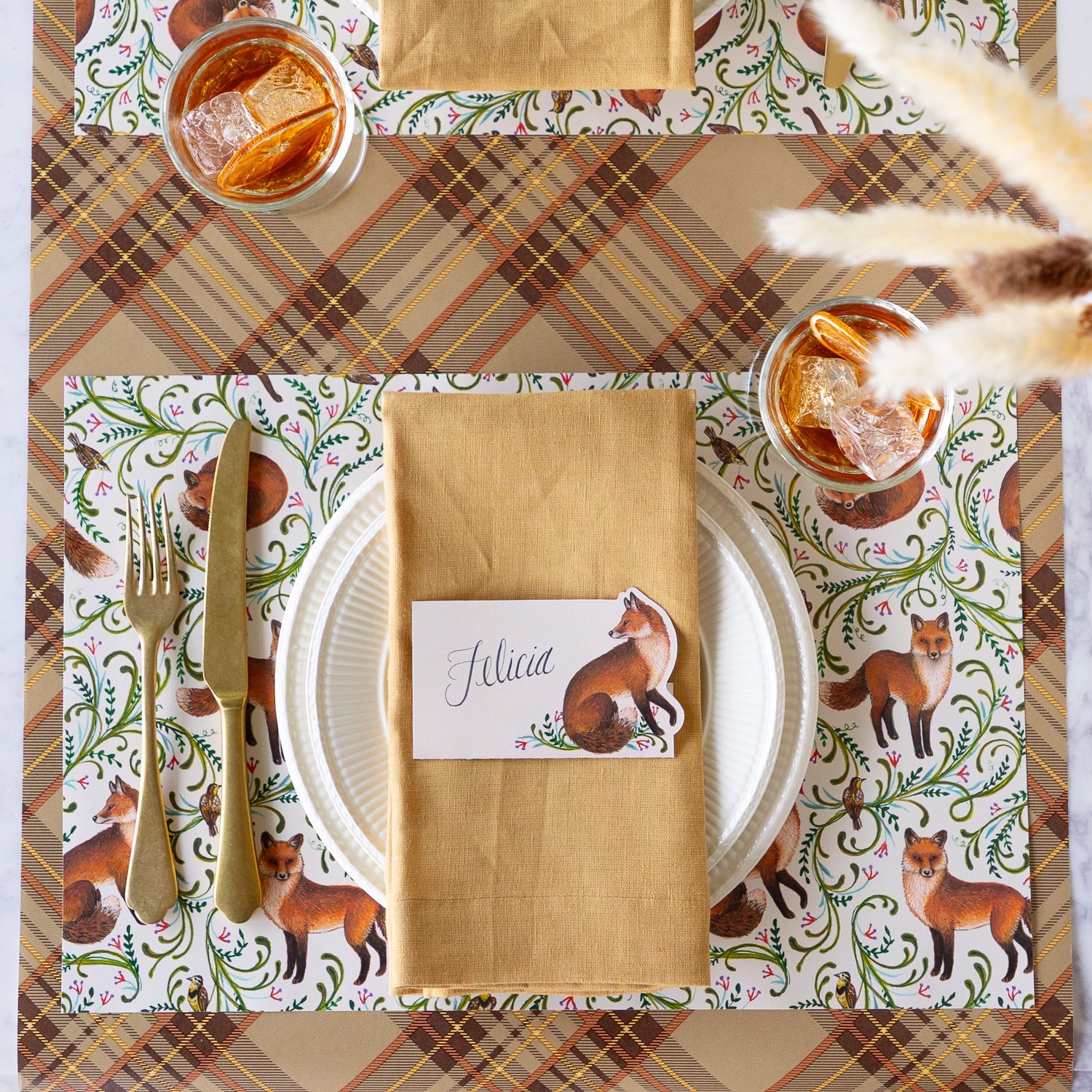 An elegant place setting featuring the Fanciful Foxes Placemat, Mepra Gold fork and knife, white dinner plates with a tan napkin and Fanciful Fox Place card on top, and Autumn Plaid Runner underneath.