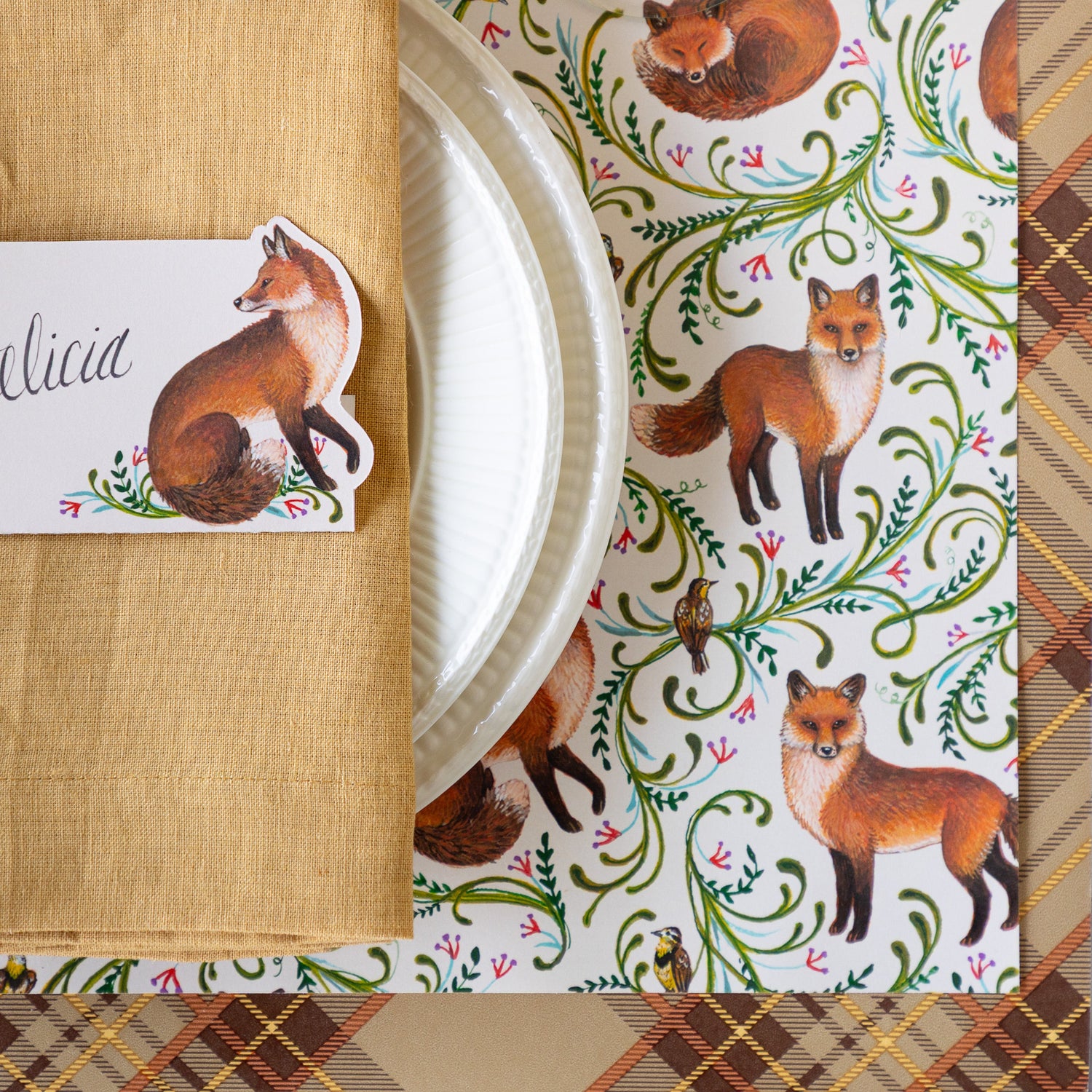 A close up of the Fanciful Fox Placemat on the Autumn Plaid Runner with white dinnerware, a tan napkin and Fanciful Fox Place Card on top.