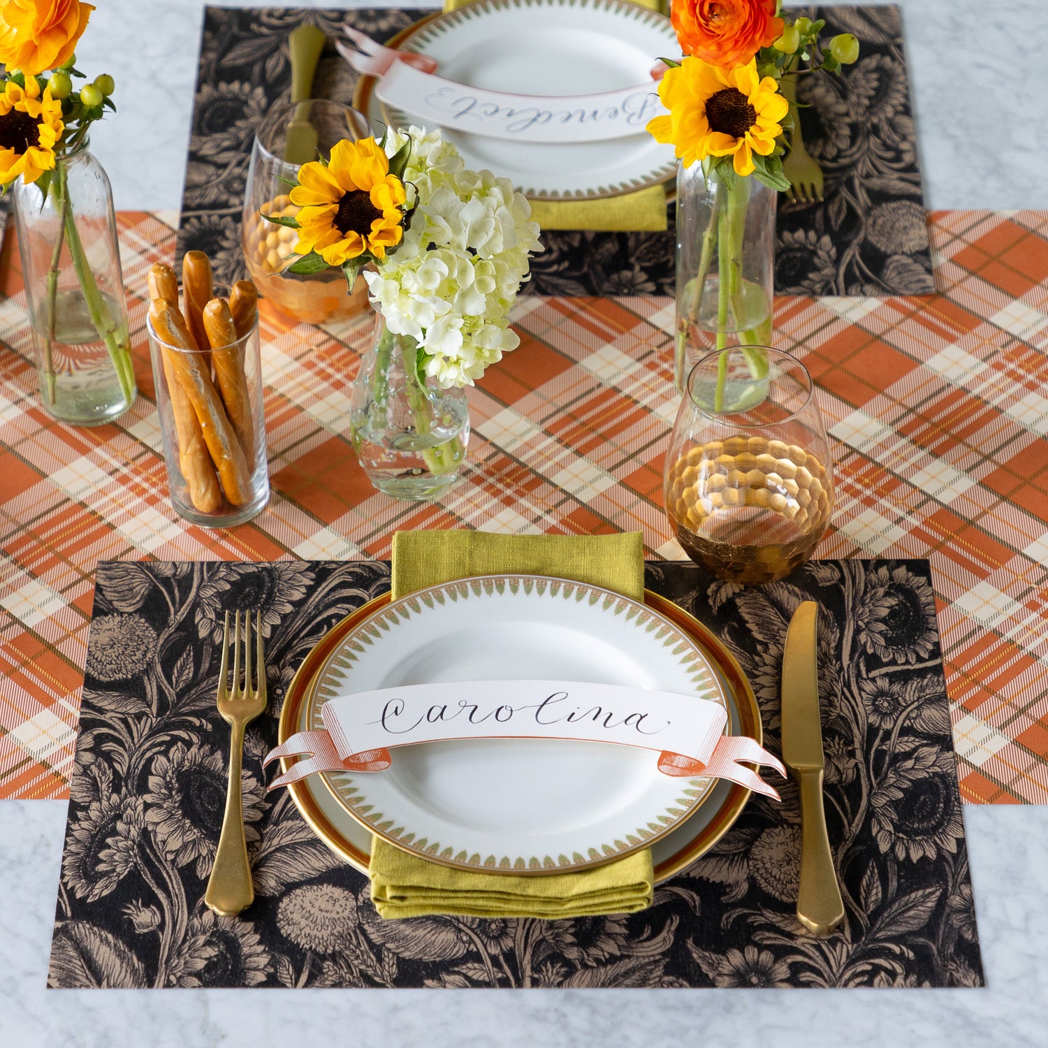 A table setting for two with fresh flowers and a cup of breadsticks, featuring the Sunflower Field Placemats, green napkins, gold flatware, vintage dinnerware with the Orange Classic Banner Table Accents atop, and Orange Plaid Runner underneath.