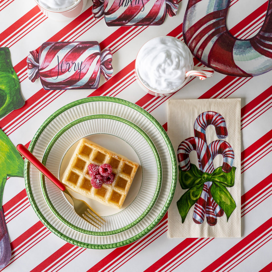 The Candy Cane Guest Napkin next to green trimmed plates with a waffle topped with raspberries, a cup filled with whip cream and a candy cane, and the Christmas Candy Cane Place Card, on the Peppermint Stripe Runner.