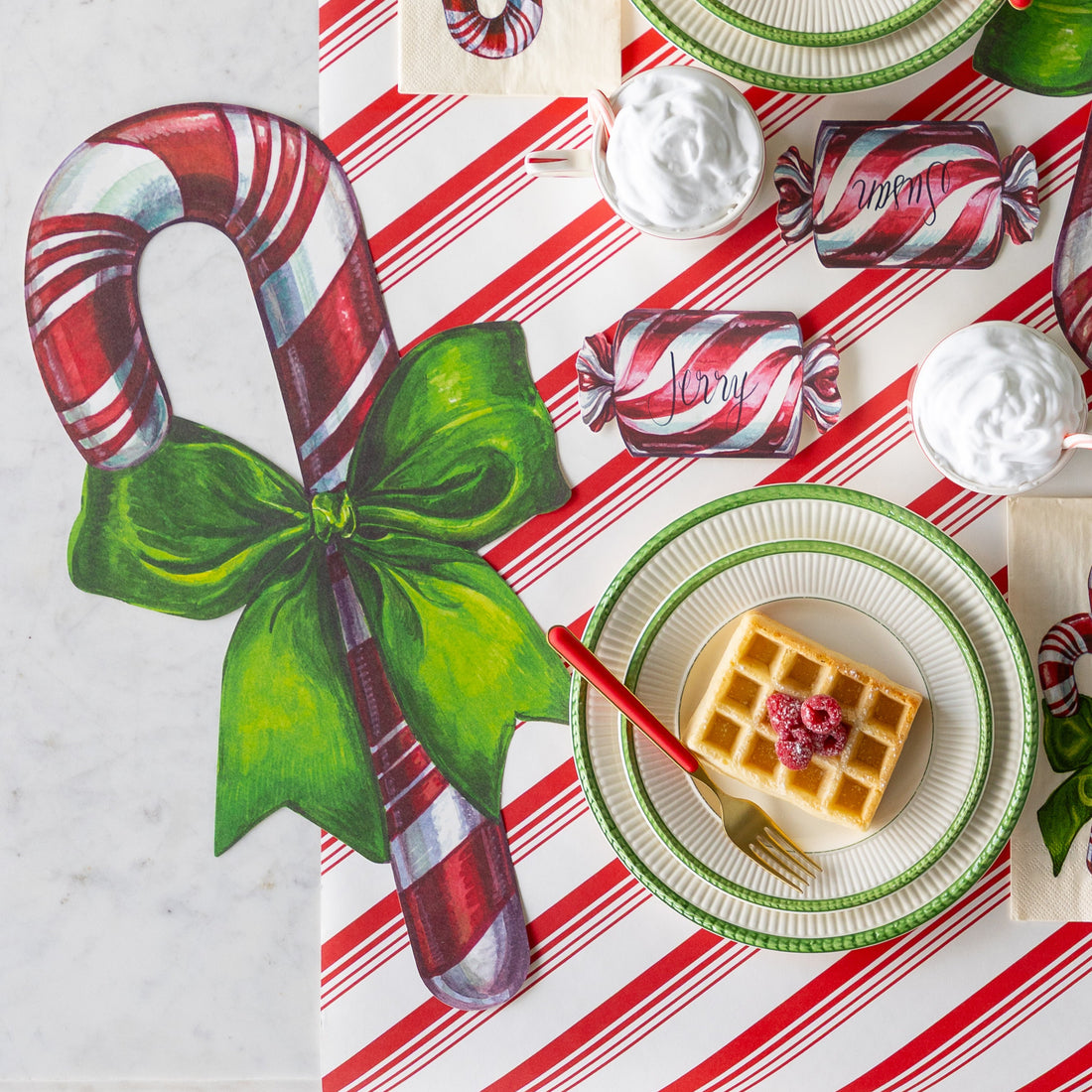 A place setting featuring the Die-cut Candy Cane Placemat next to a plate of waffles with a red and gold fork on top, Christmas Candy Place Card reading "Jerry" and Peppermint Stripe Runner underneath.
