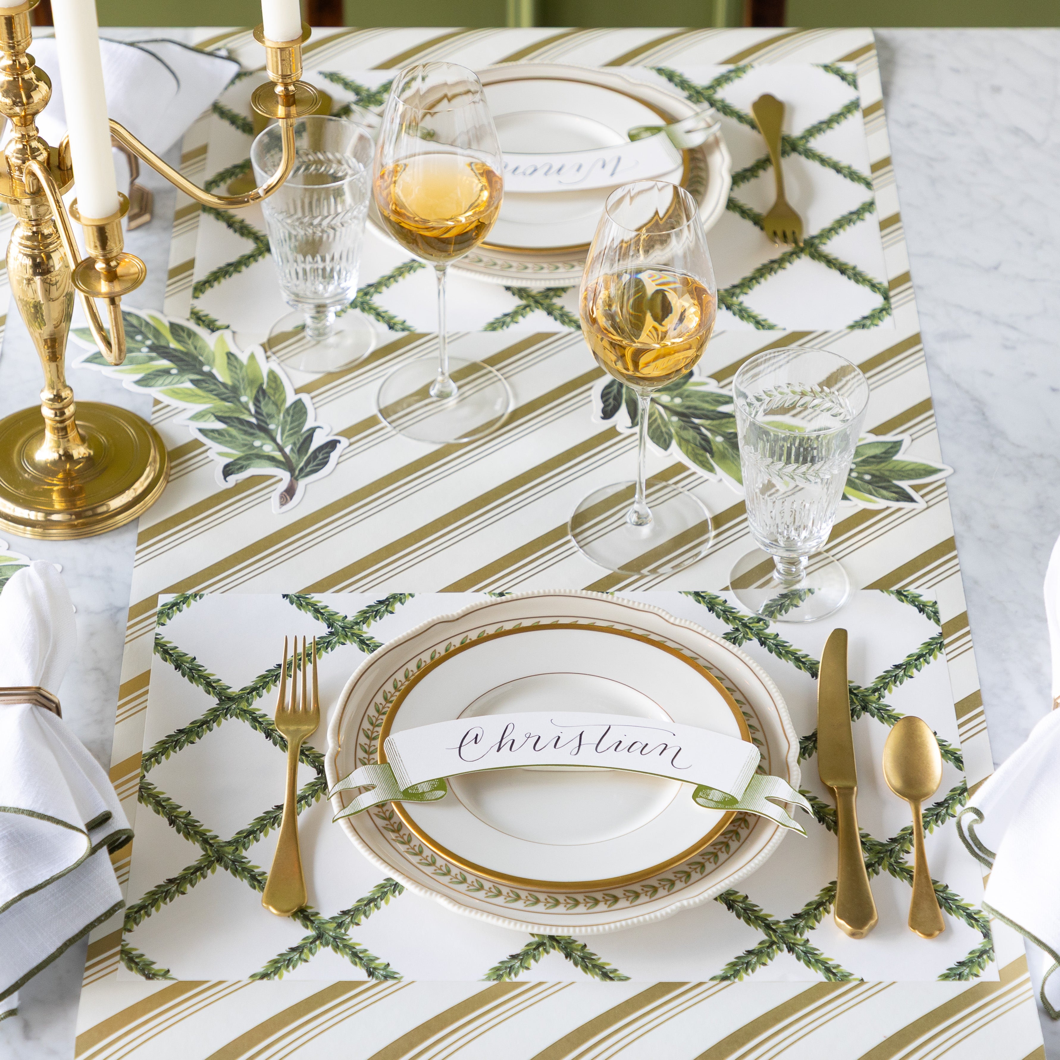 A formal table setting with gold cutlery, wine glasses, vintage plates with the Classic Moss Banner Table Accent atop, Laurel Table Accents scattered on the table, and Laurel Wreath Placemats on the Gold Peppermint Stripe Runner.