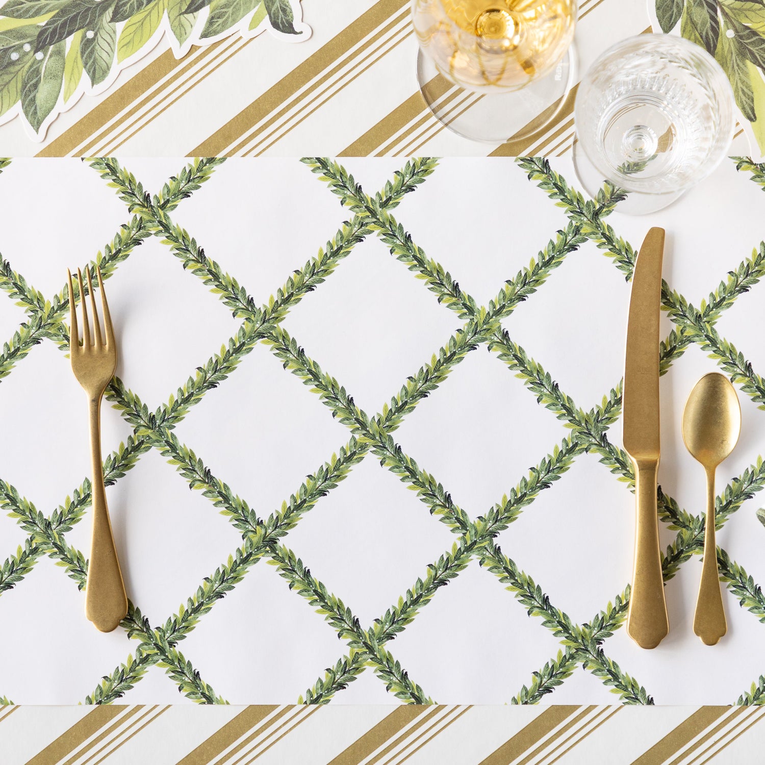 The Laurel Wreath Placemat with gold flatware on top, and a wine and water glass, on top of the Gold Peppermint Stripe Runner.