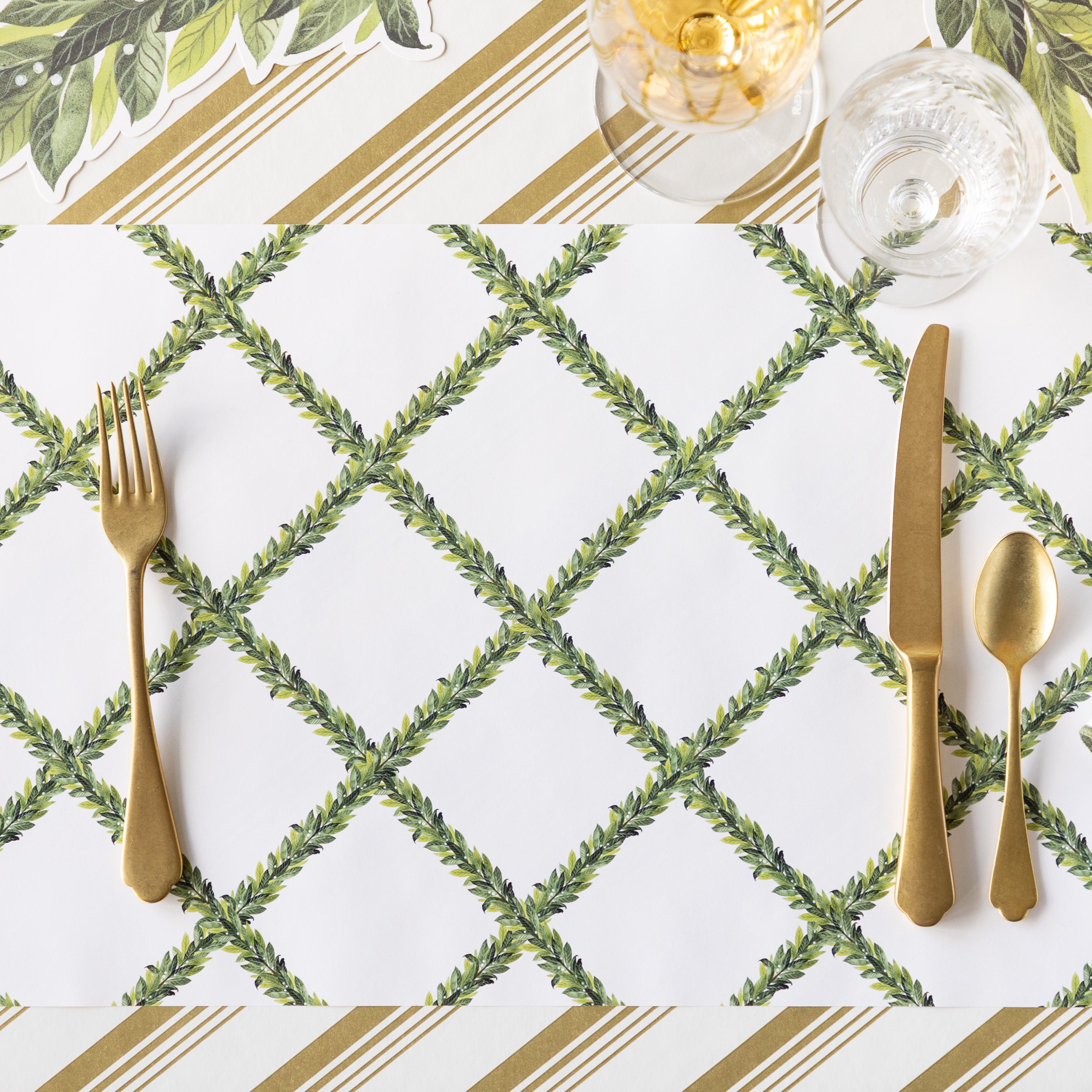 The Laurel Wreath Placemat with gold flatware on top, and a wine and water glass, on top of the Gold Peppermint Stripe Runner.