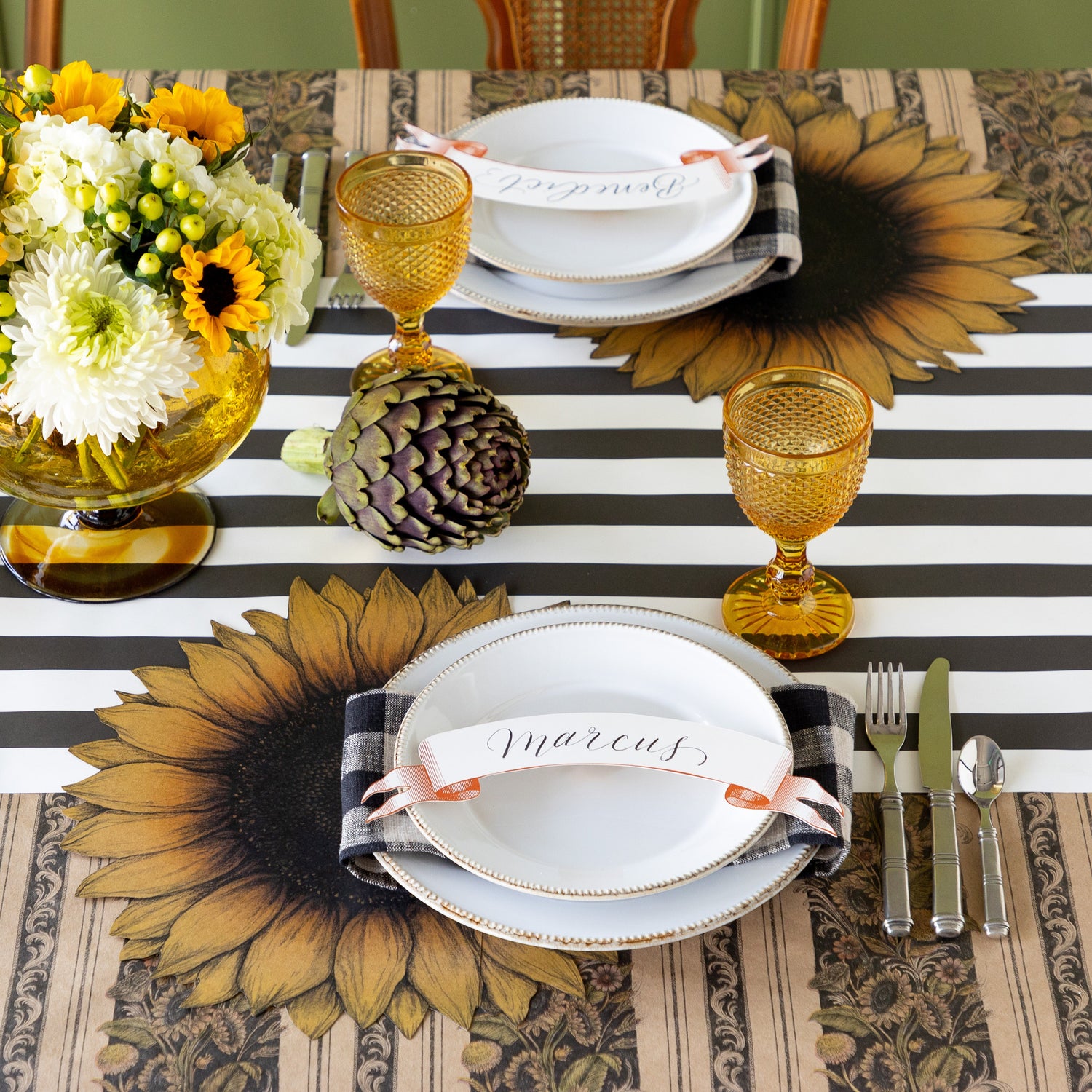 A table setting with fresh flowers centerpiece and an artichoke featuring the Die-cut Vintage Sunflower Placemat under white dinner plates with the Classic Orange Banner Table Accent atop, Vintage Black Sunflower Stripe Runner rolled vertically and Black Classic Stripe Runner rolled out horizontally underneath.