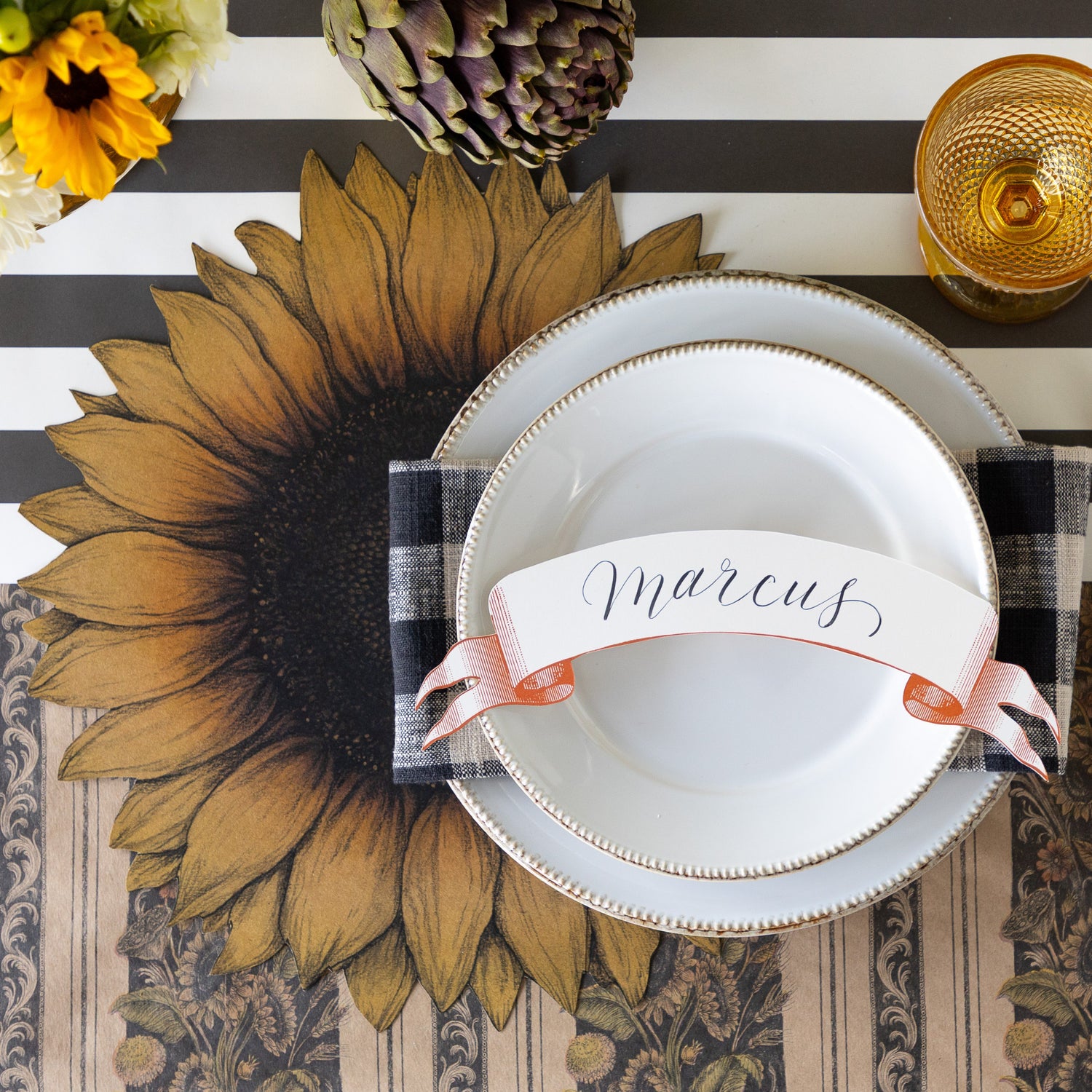 A place setting with fresh flowers and an artichoke, featuring the Die-cut Vintage Sunflower Placemat, a black checkered napkin, two white plates with the Classic Orange Banner Table Accent reading &quot;Marcus&quot; atop, Vintage Black Sunflower Runner rolled out vertically and Black Classic Stripe Runner horizontally underneath.