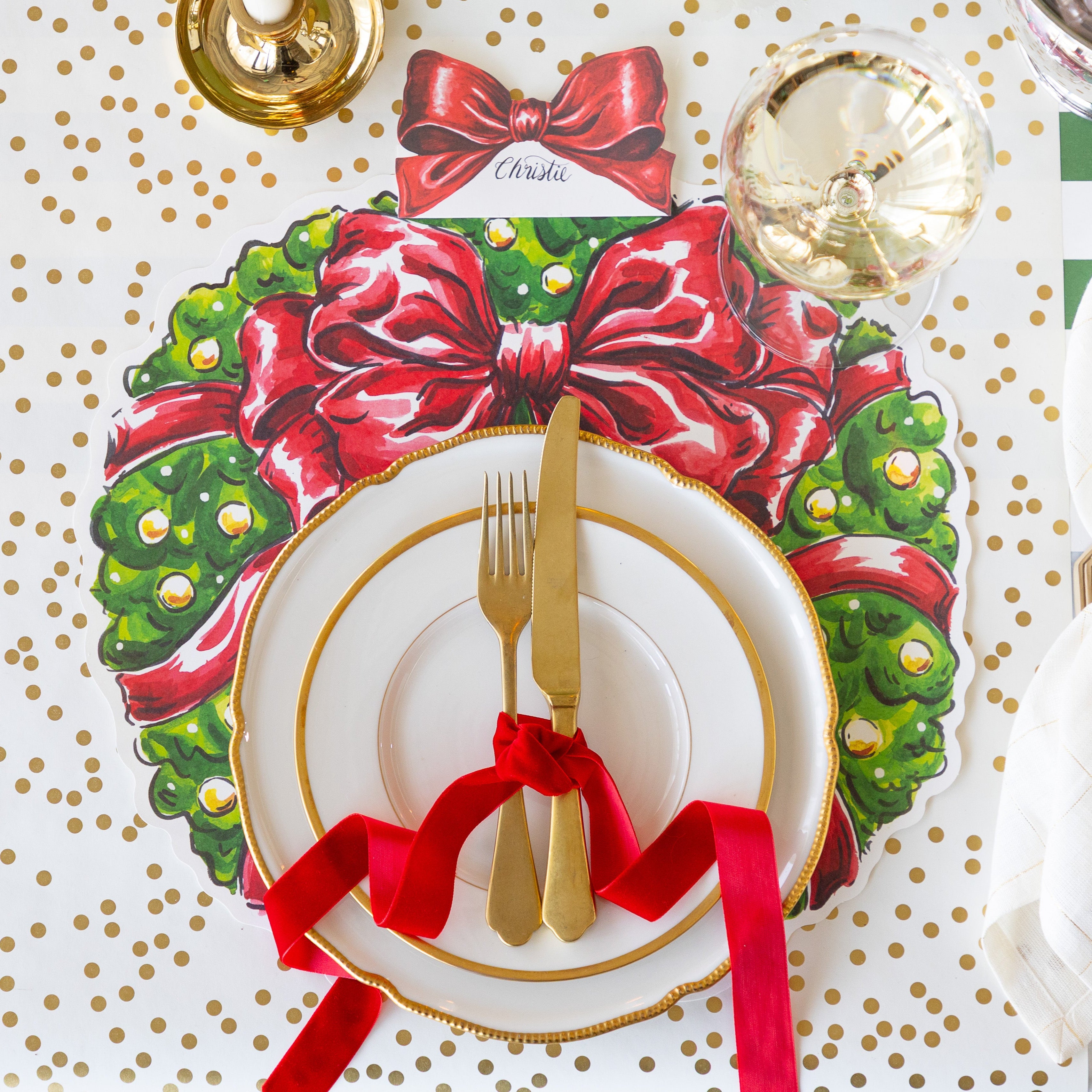 A place setting featuring the Die-cut Holiday Wreath Placemat with gold rimmed plates and a gold fork and knife tied in a red ribbon, atop, Bow Place Card reading "Christie, and a glass of wine on the Gold Confetti Runner.