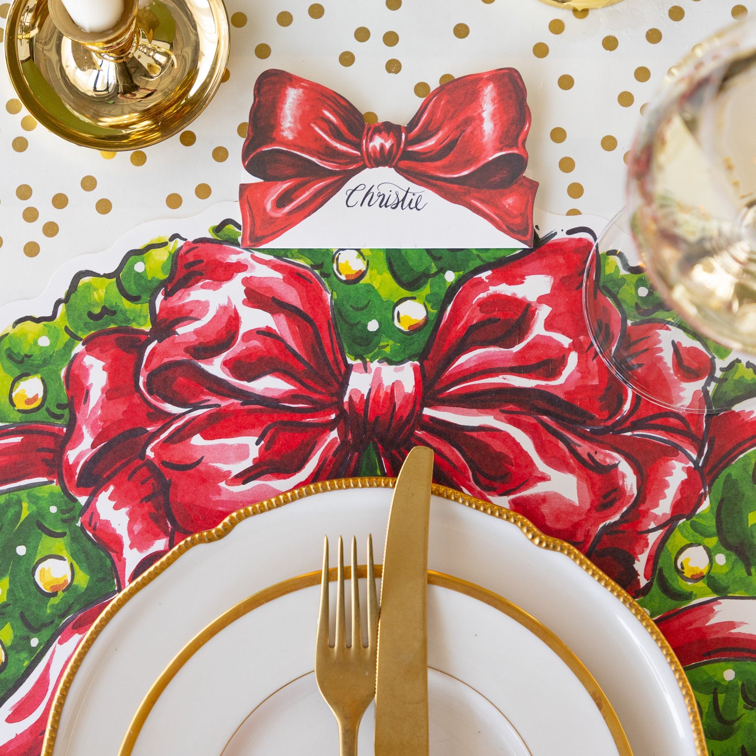 The Bow Place Card reading &quot;Christie&quot; behind a place setting with the Die-cut Holiday Wreath Placemat, gold rimmed dinner plates and a gold fork and knife on the Gold Confetti Runner.