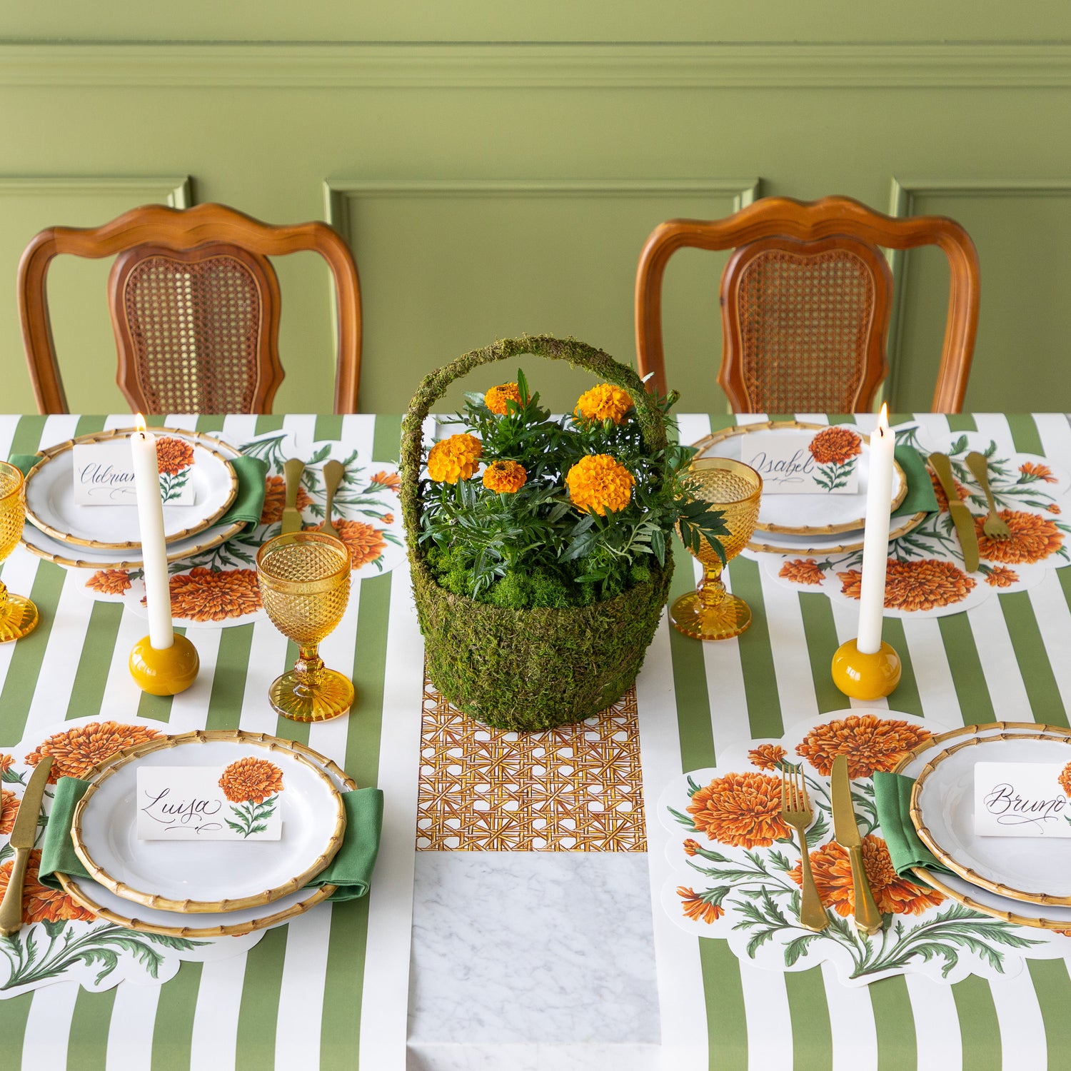A dining table is set with Die-cut Marigold Bouquet Placemats, bamboo plates with Marigold Bloom Place Cards on top, yellow glasses, lit candles, a centerpiece moss basket of Marigolds and Moss Classic Stripe Runners vertically under the place settings with the Rattan Weave Runner running horizontally underneath it.