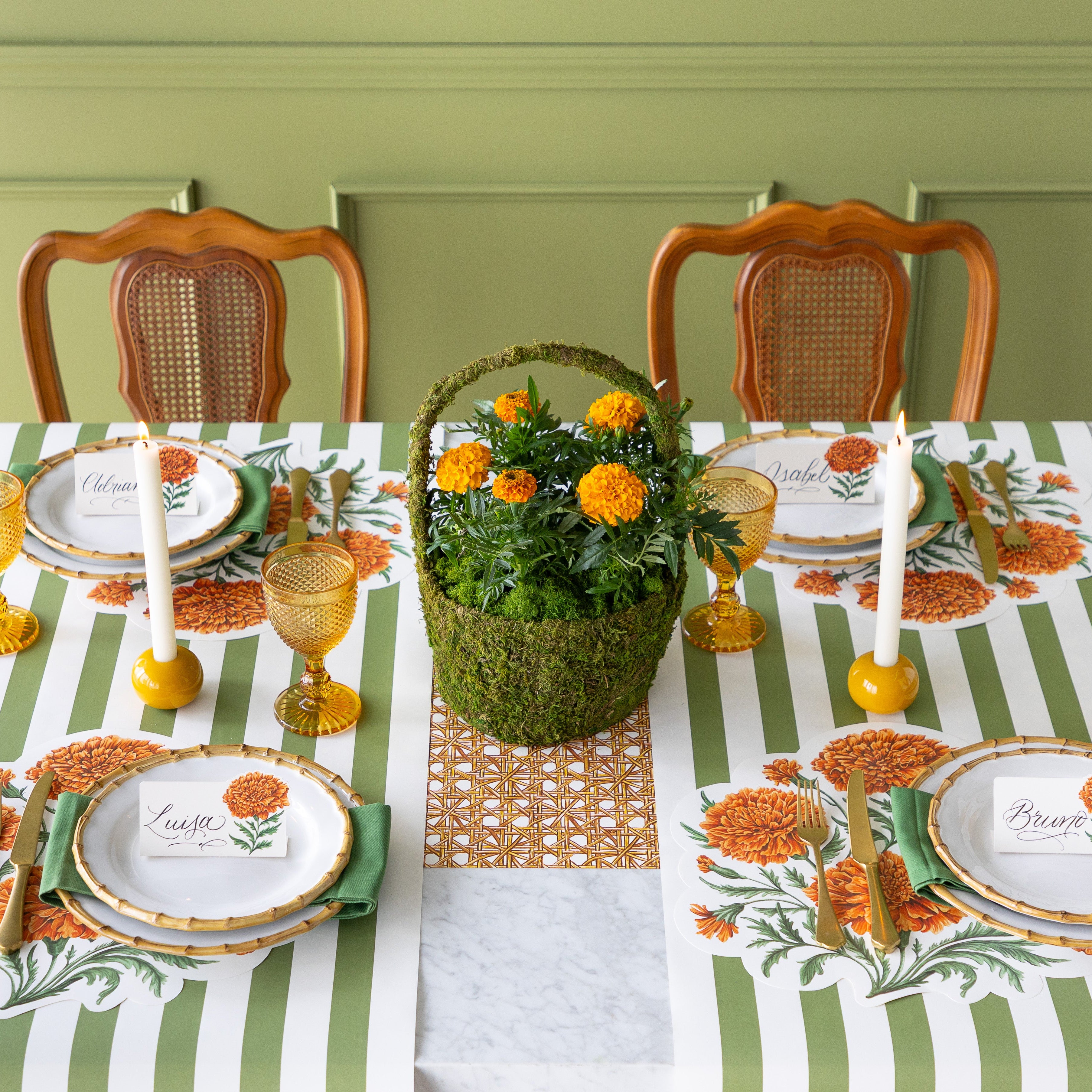 A dining table is set with Die-cut Marigold Bouquet Placemats, bamboo plates with Marigold Bloom Place Cards on top, yellow glasses, lit candles, a centerpiece moss basket of Marigolds and Moss Classic Stripe Runners vertically under the place settings with the Rattan Weave Runner running horizontally underneath it.