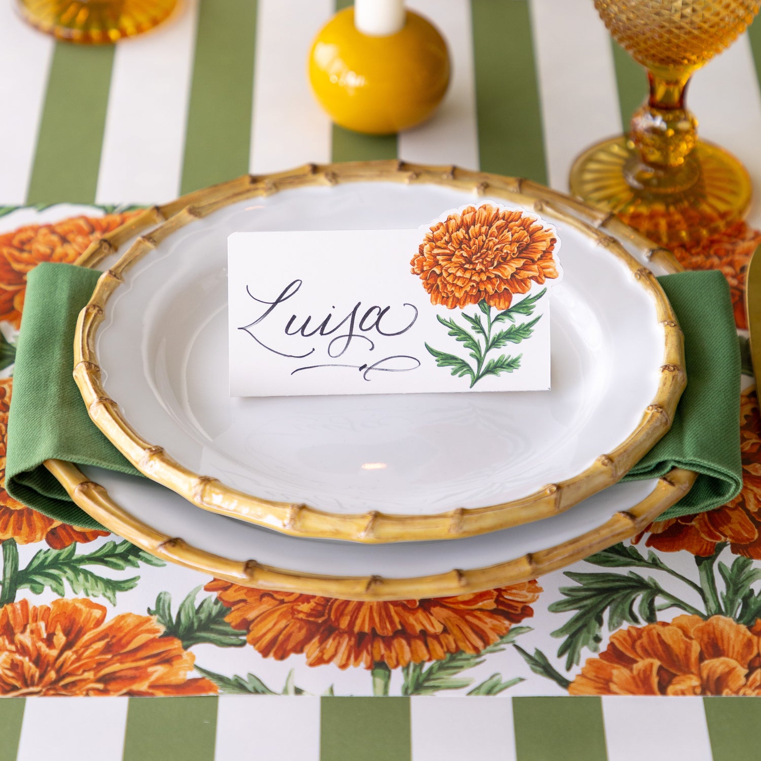 A place setting featuring the Marigold Bloom Place Card reading &quot;Luisa&quot; atop bamboo plates with the Marigold Harvest Placemat and Moss Classic Stripe Runner underneath.