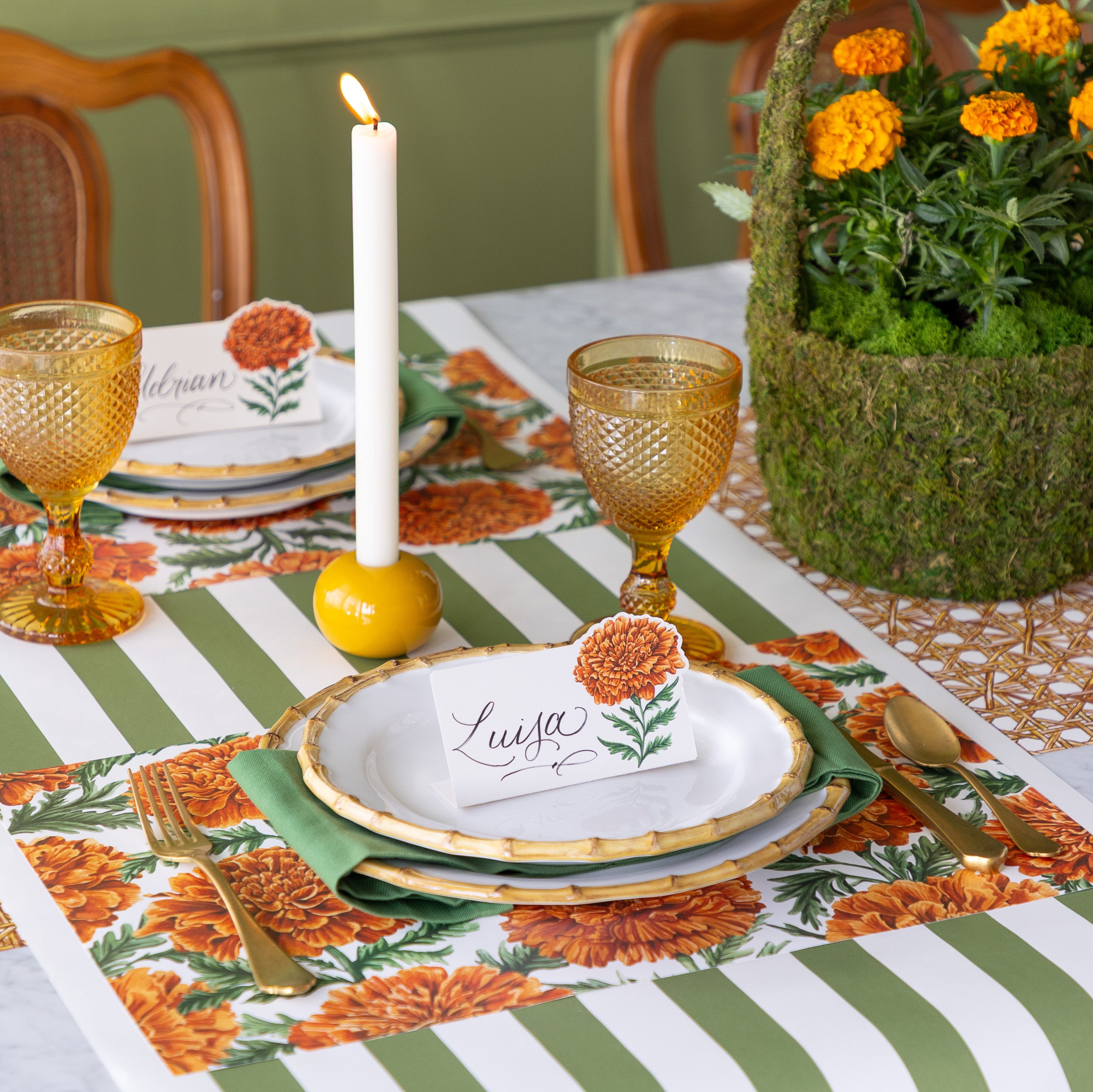 A set table with a lit candle and a Moss basket centerpiece filled with Marigolds, featuring the Marigold Harvest Placemat under bamboo dinnerware with the Marigold Bloom Place Card reading &quot;Luis&quot; atop, Moss Classic Stripe Runner vertically on the table and Rattan Reave Runner horizontally underneath.