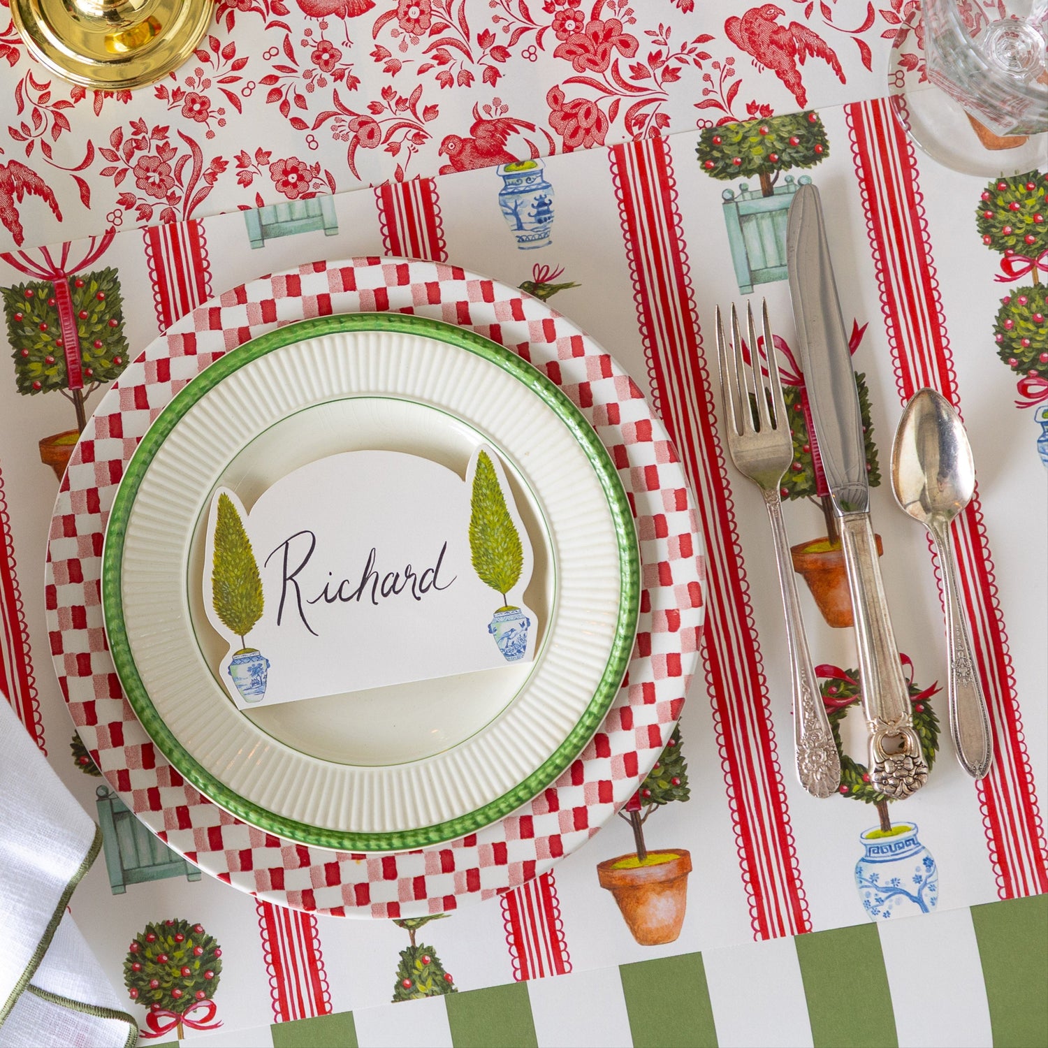 An elegant place setting featuring the Holiday Topiary Placemat, dinner plates with the Topiary Place Card on top, Moss Classic Stripe Runner running vertically and Red Regal Peacock Runner running horizontally underneath.