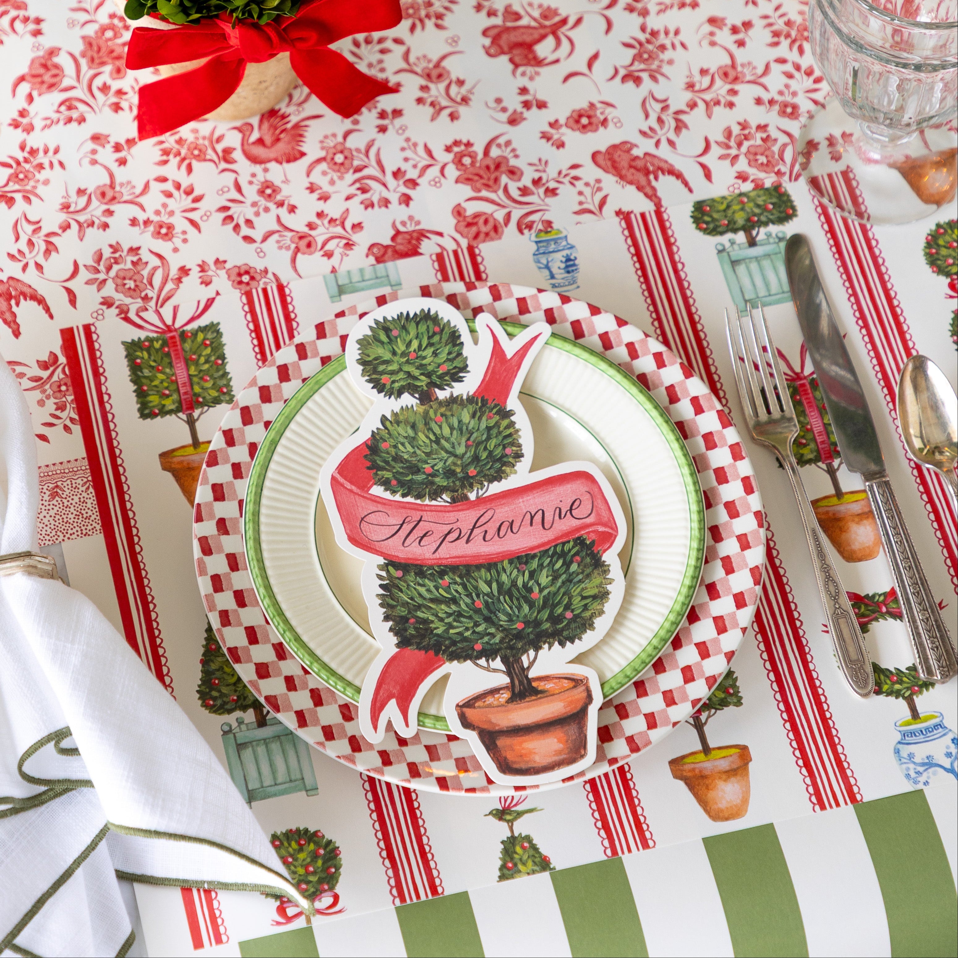An elegant place setting featuring the Holiday Topiary Placemat, dinner plates with the Holiday Topiary Table Accent on top, Moss Classic Stripe Runner running vertically and Red Regal Peacock Runner running horizontally underneath.