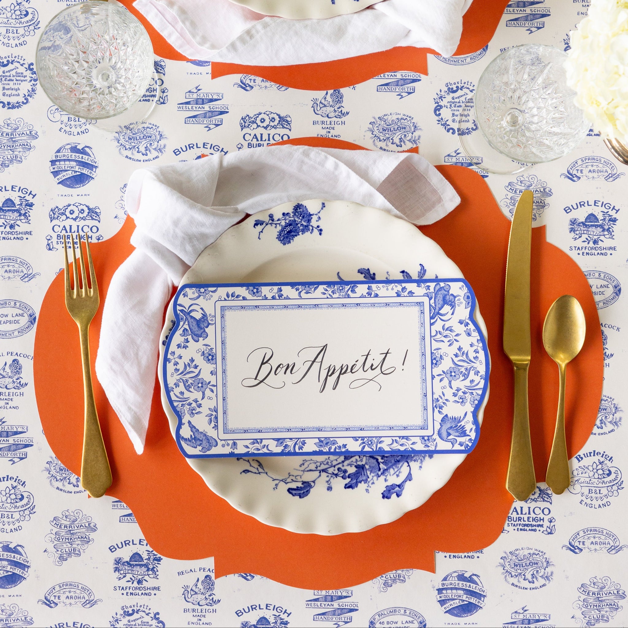 A place setting featuring the Die-cut Orange French Frame Placemat with a knotted napkin, floral plate, Blue Regal Peacock Table Accent reading &quot;Bon Appetit!&quot;and gold flatware atop, on the Burleigh Heritage Runner.