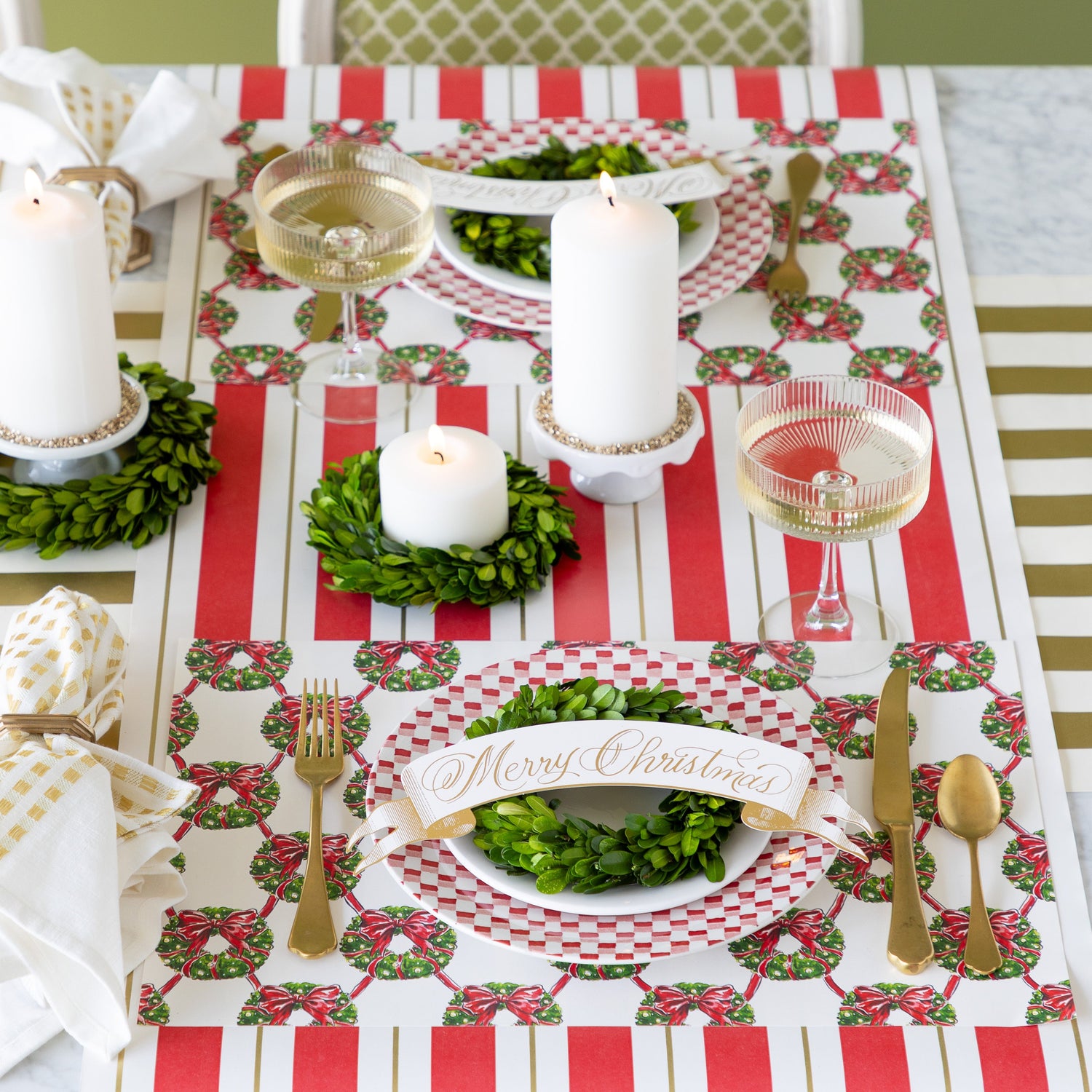 A table setting with lit candles and boxwood wreaths around them, Holiday Wreath Lattice Placemats, red-and-white checkered plates with boxwood wreaths and Merry Christmas Banner Table Accents atop, gold flatware, and champagne coupe glasses on the Red &amp; Gold Awning Stripe and Gold Classic Stripe Runners.