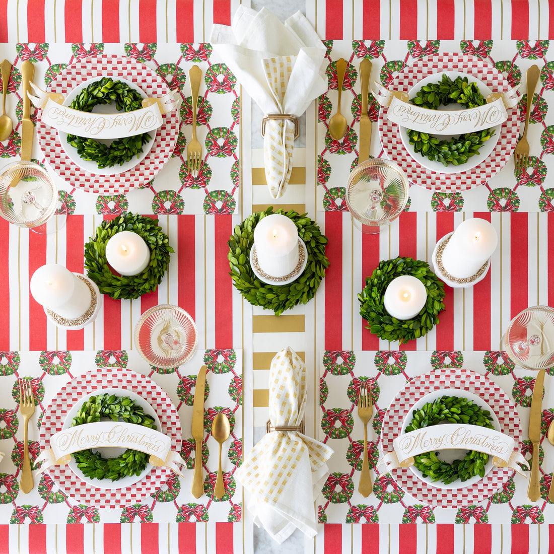 A festive table setting featuring Holiday Wreath Lattice Placemats, red-and-white plates adorned with  wreaths and Merry Christmas Banner Table Accents, candles, and gold accents on the Antique Red Striped Runner.