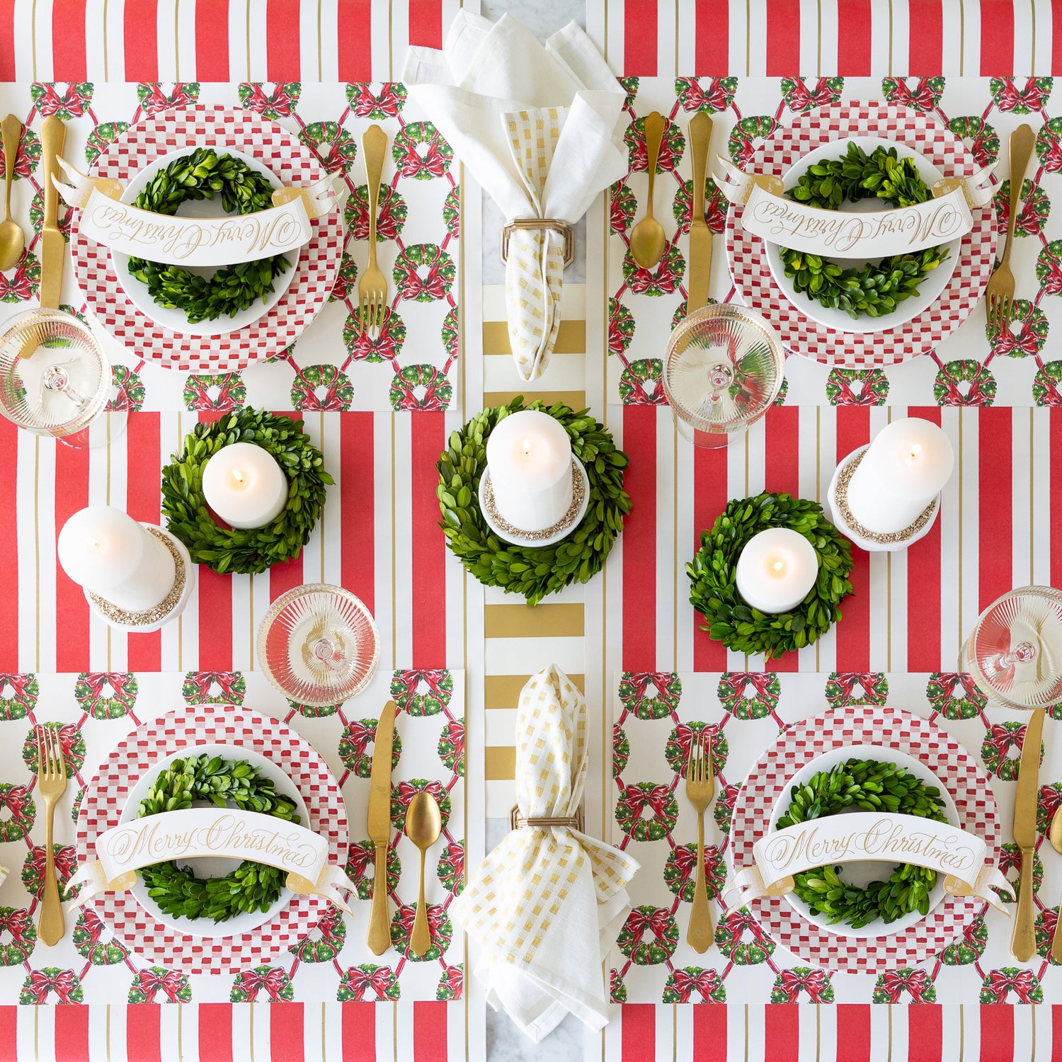 A festive table setting featuring Holiday Wreath Lattice Placemats, red-and-white plates adorned with  wreaths and Merry Christmas Banner Table Accents, candles, and gold accents on the Antique Red Striped Runner.