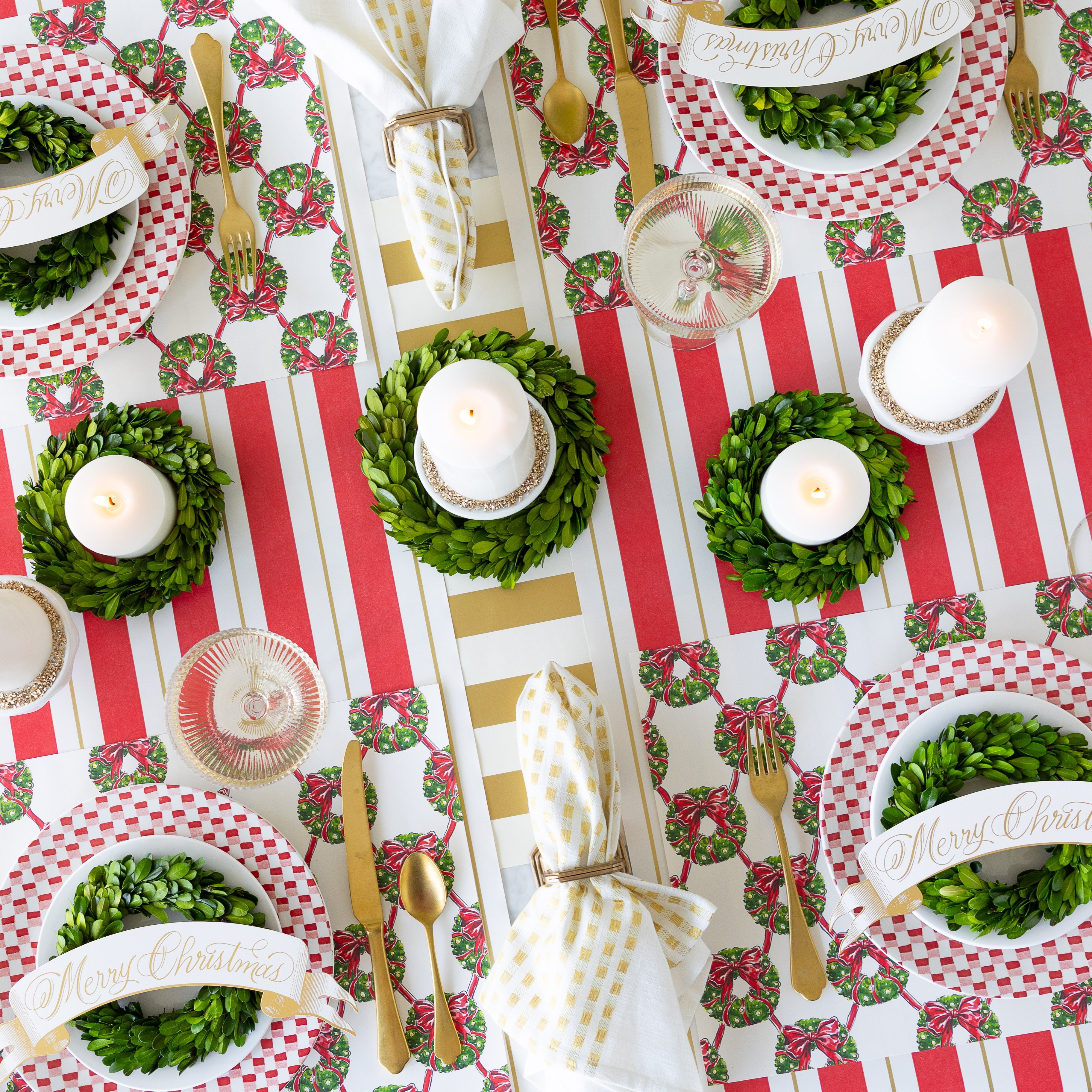 An elegant Christmas table setting with candles, boxwood wreath, Holiday Wreath Lattice Placemats, Merry Christmas Banner Table Accents, and napkins in a holder on the Red &amp; Gold Awning Stripe and Gold Classic Stripe Runners.