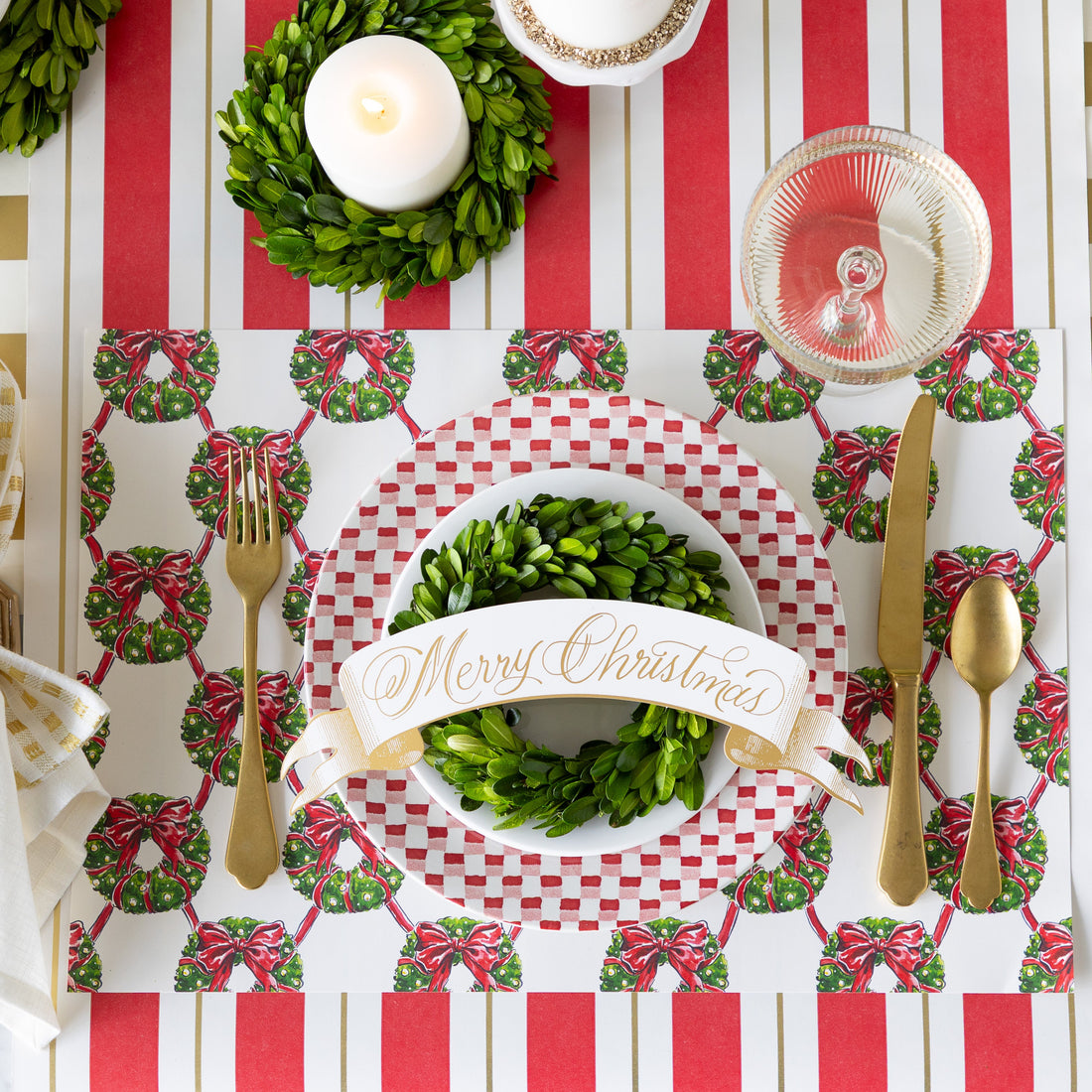 A place setting featuring the Holiday Wreath Lattice Placemat, a red-and-white checkered plate with a white plate, boxwood wreath and Merry Christmas Banner Table Accent atop, gold flatware, and coupe glass on the Red &amp; Gold Awning Stripe Runner.