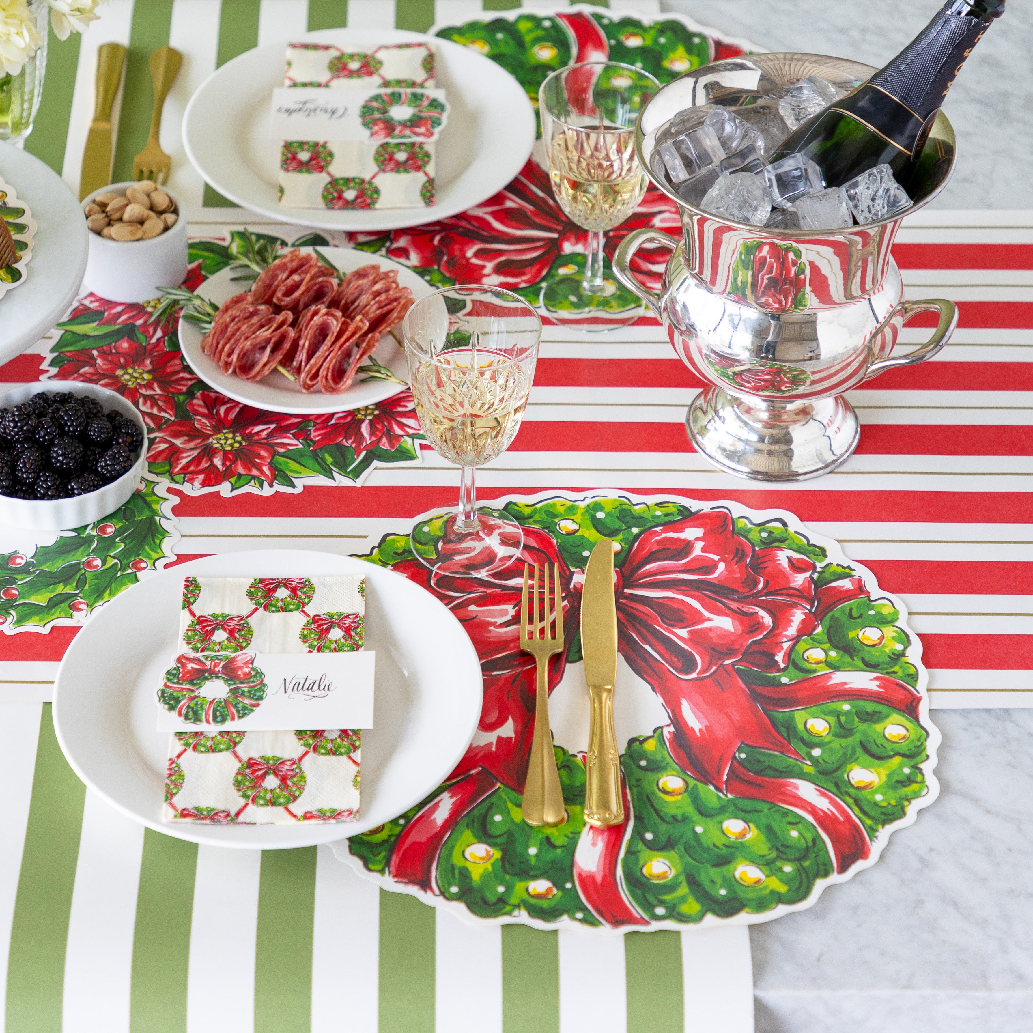 A festive table setting with charcuterie and champagne on ice, featuring the Die-cut Holiday Wreath Placemat, a plate with the Holiday Wreath Guest Napkin and Holiday Wreath Place Card atop, and gold cutlery on the Red &amp; Gold Awning Stripe Runner and Moss Classic Stripe Runner.