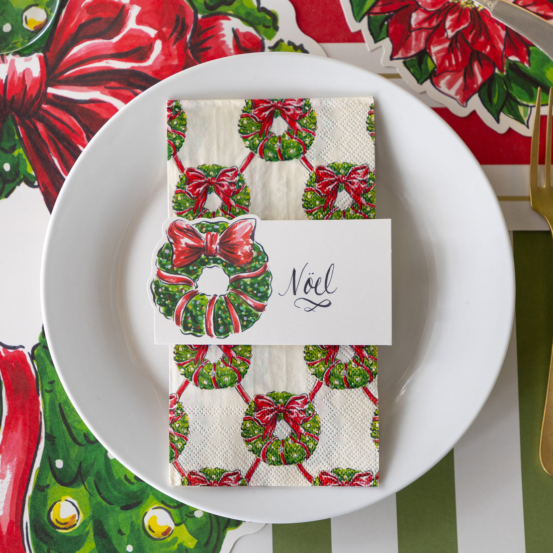 A place setting with the Holiday Wreath Place Card reading "Noel", atop the Holiday Wreath Guest Napkin and white plate, on the Die-cut Holiday Wreath Placemat.
