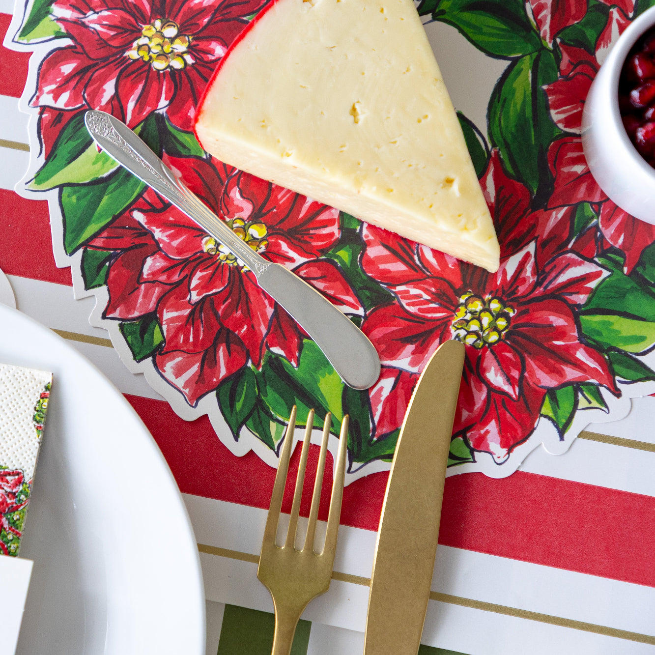 A Holiday Wreath Serving Paper behind a place setting with a sliced block of cheese and butter spreader atop.