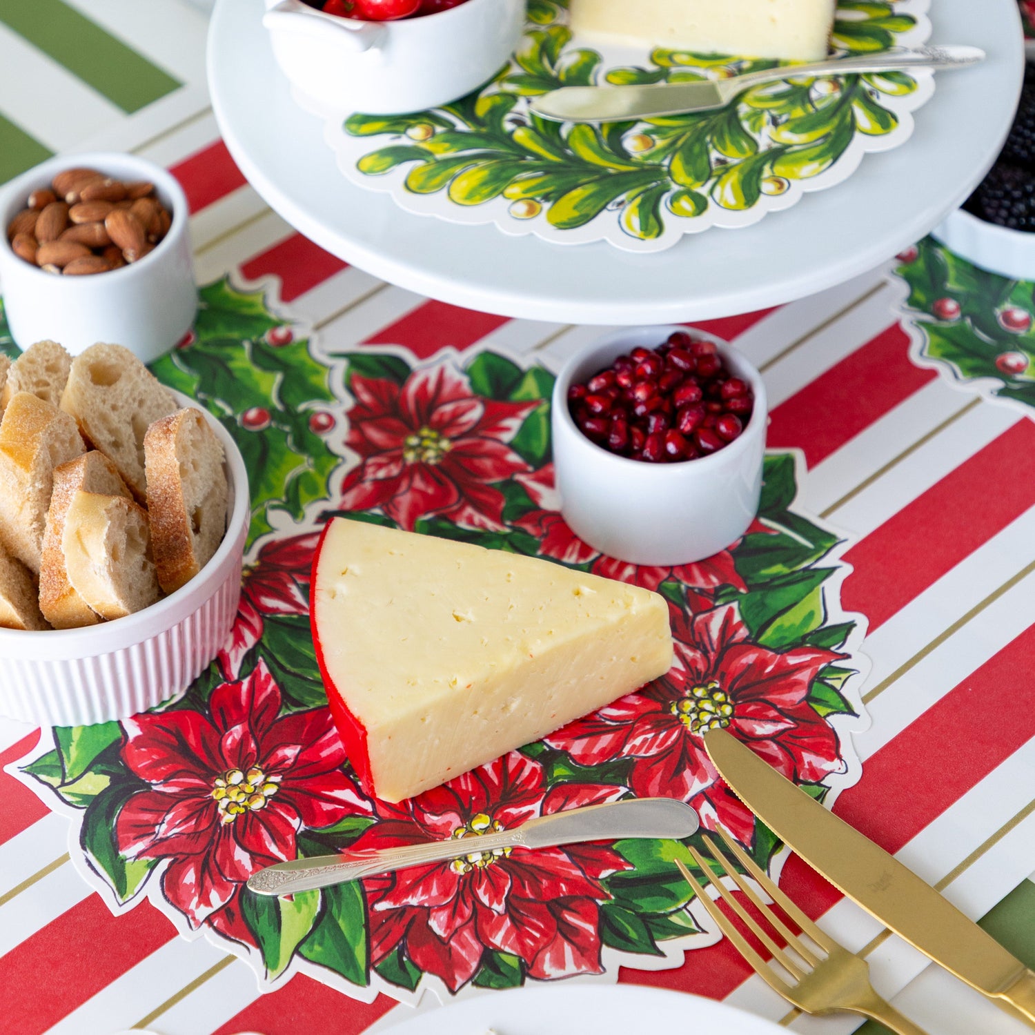 A table setting with a Holiday Wreath Serving Paper below a block of cheese, bowl of sliced bread and pomegranate seeds, and a butter spreader.