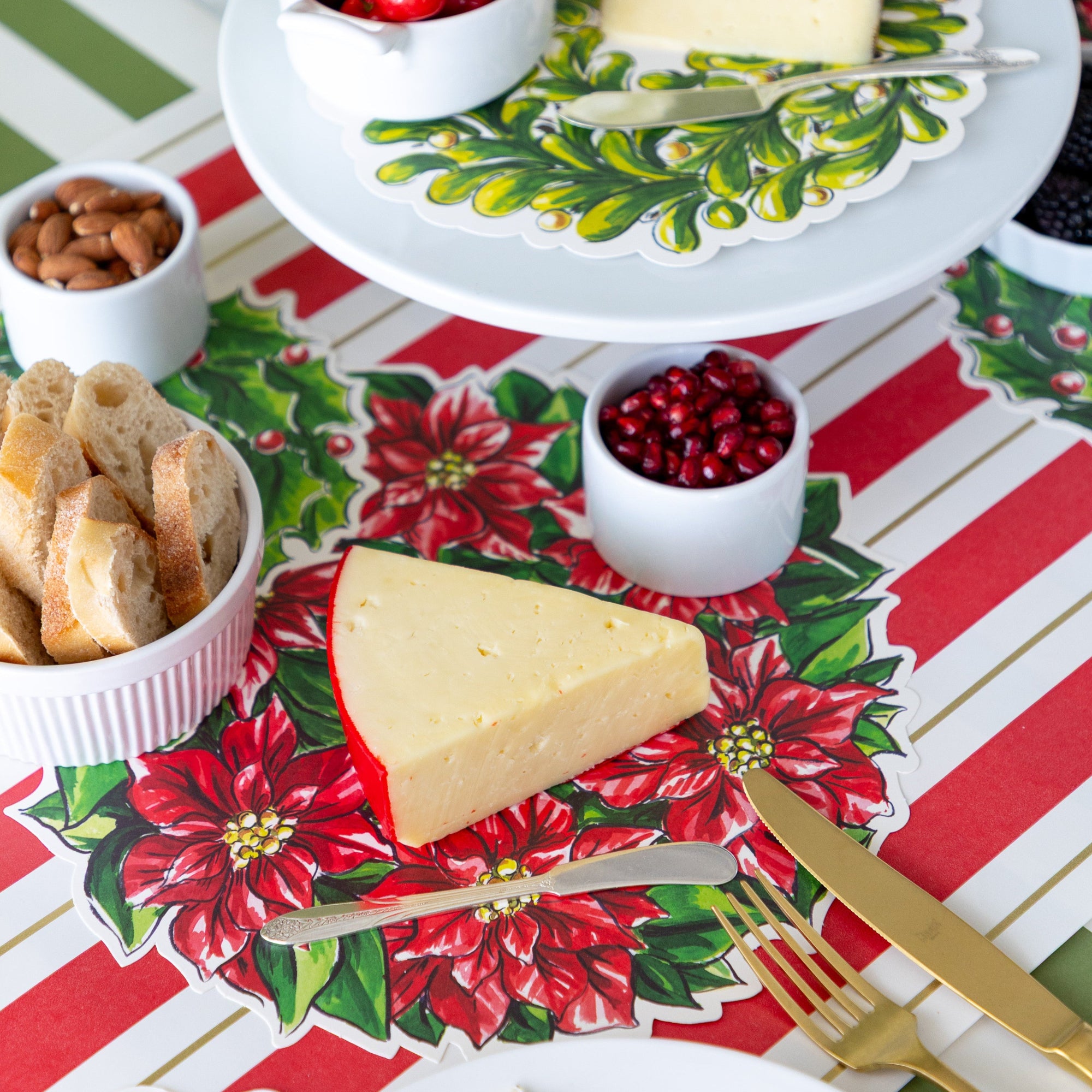 A table setting with a Holiday Wreath Serving Paper below a block of cheese, bowl of sliced bread and pomegranate seeds, and a butter spreader.