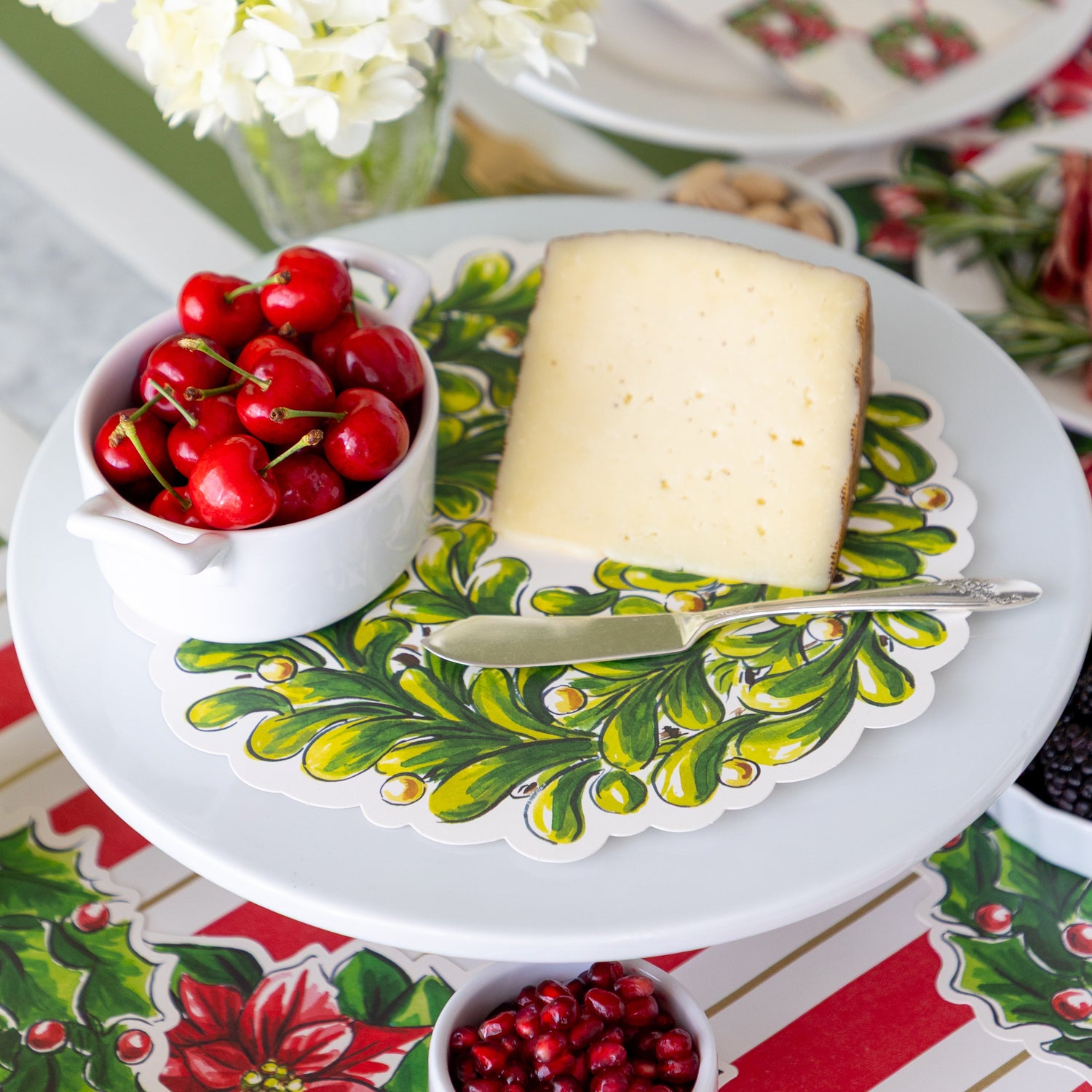 A Holiday Wreath Serving Paper on a cake stand with a bowl of cherries, a sliced block of cheese and a butter spreader atop.