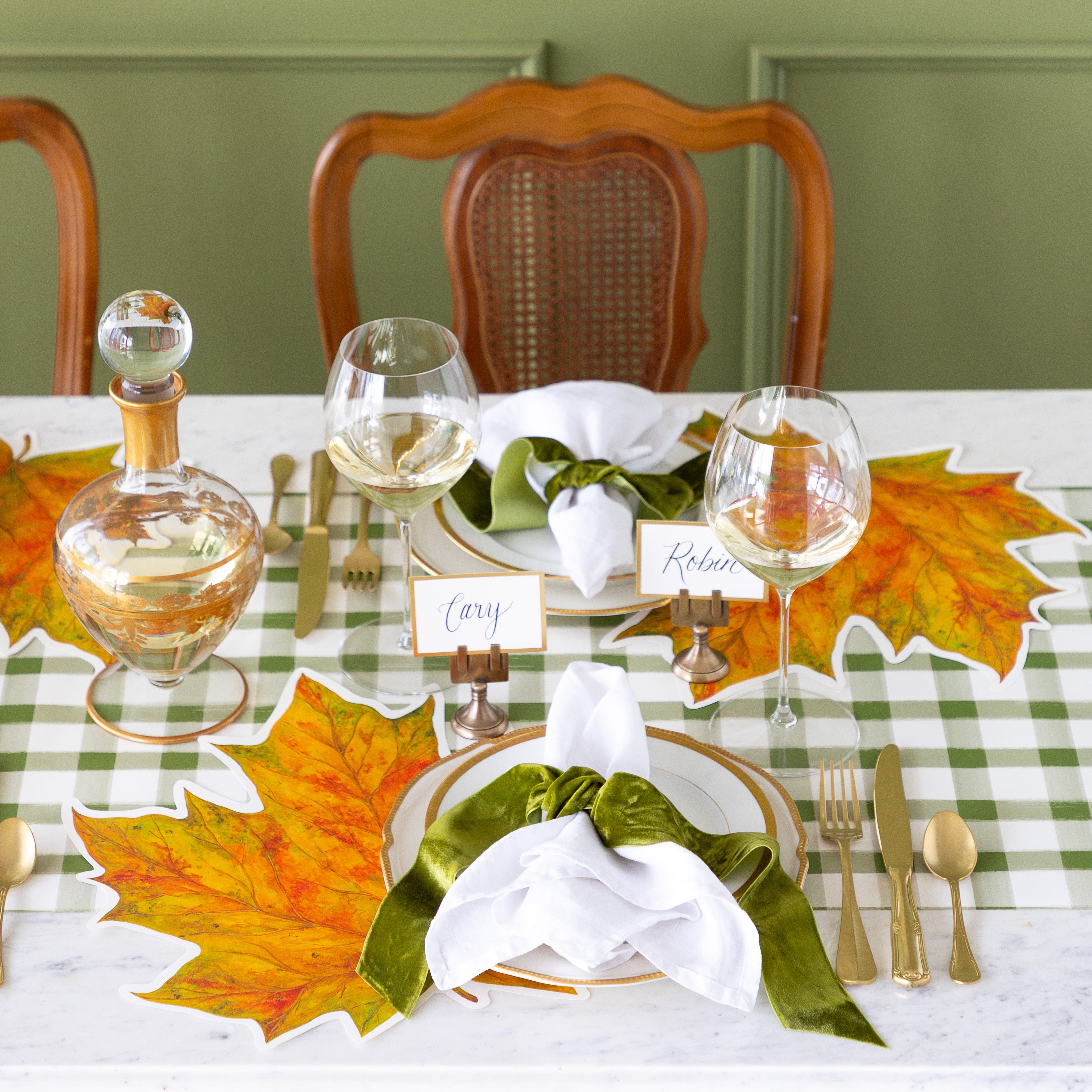 An elegant place setting featuring the Die-cut Fall Leaf Placemat, gold rimmed plates, the Gold Frame Place Card held by the Brass Place Card Holder, white napkins tied by a green velvet ribbon and the Green Painted Check Runner underneath.