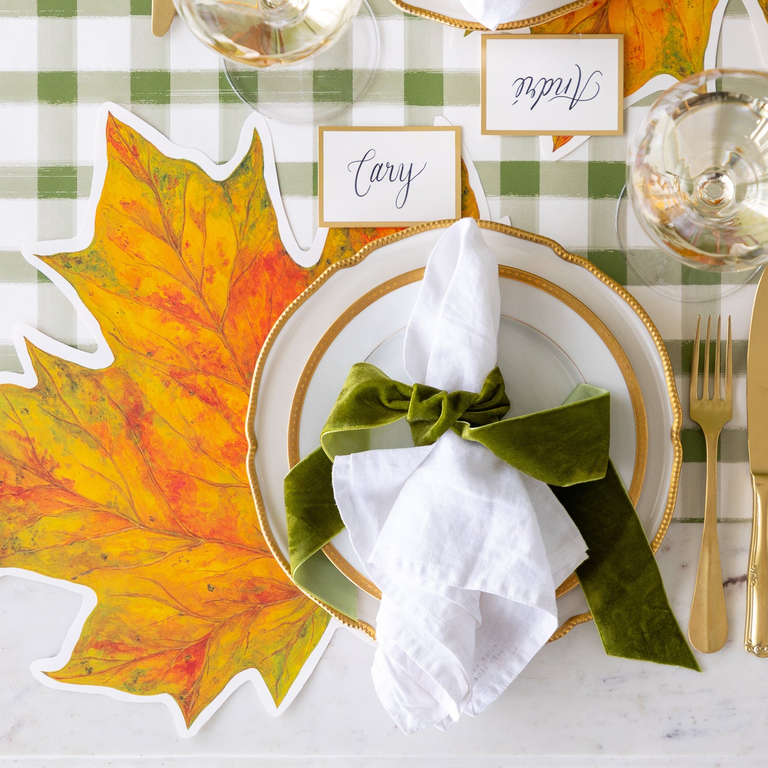 An elegant place setting featuring the Die-cut Fall Leaf Placemat, gold rimmed plates, the Gold Frame Place Card reading &quot;Cary&quot;, white napkins tied by a green velvet ribbon and the Green Painted Check Runner underneath.