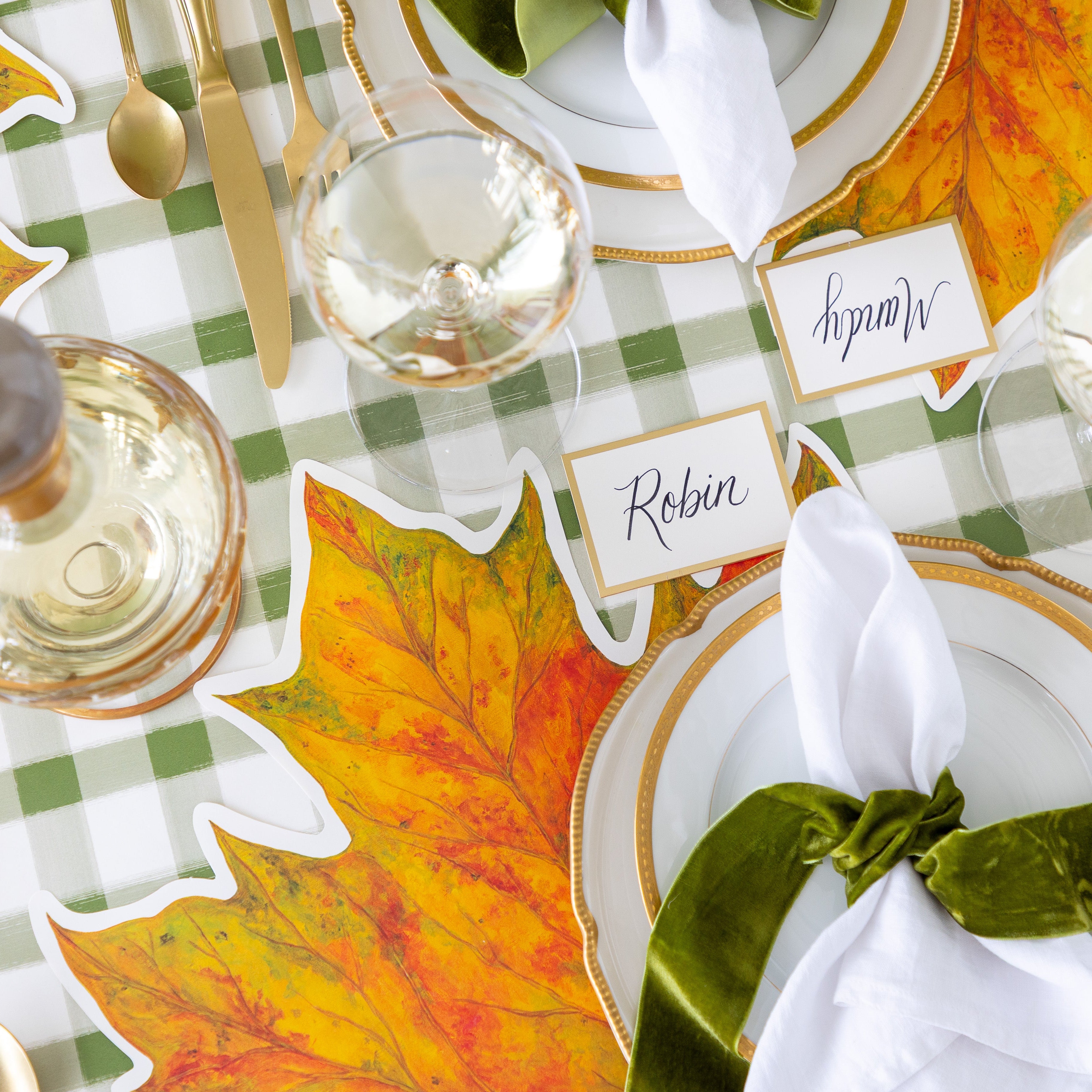An elegant place setting featuring the Die-cut Fall Leaf Placemat, gold rimmed plates, the Gold Frame Place Card reading &quot;Robin&quot;, white napkins tied by a green velvet ribbon and the Green Painted Check Runner underneath.