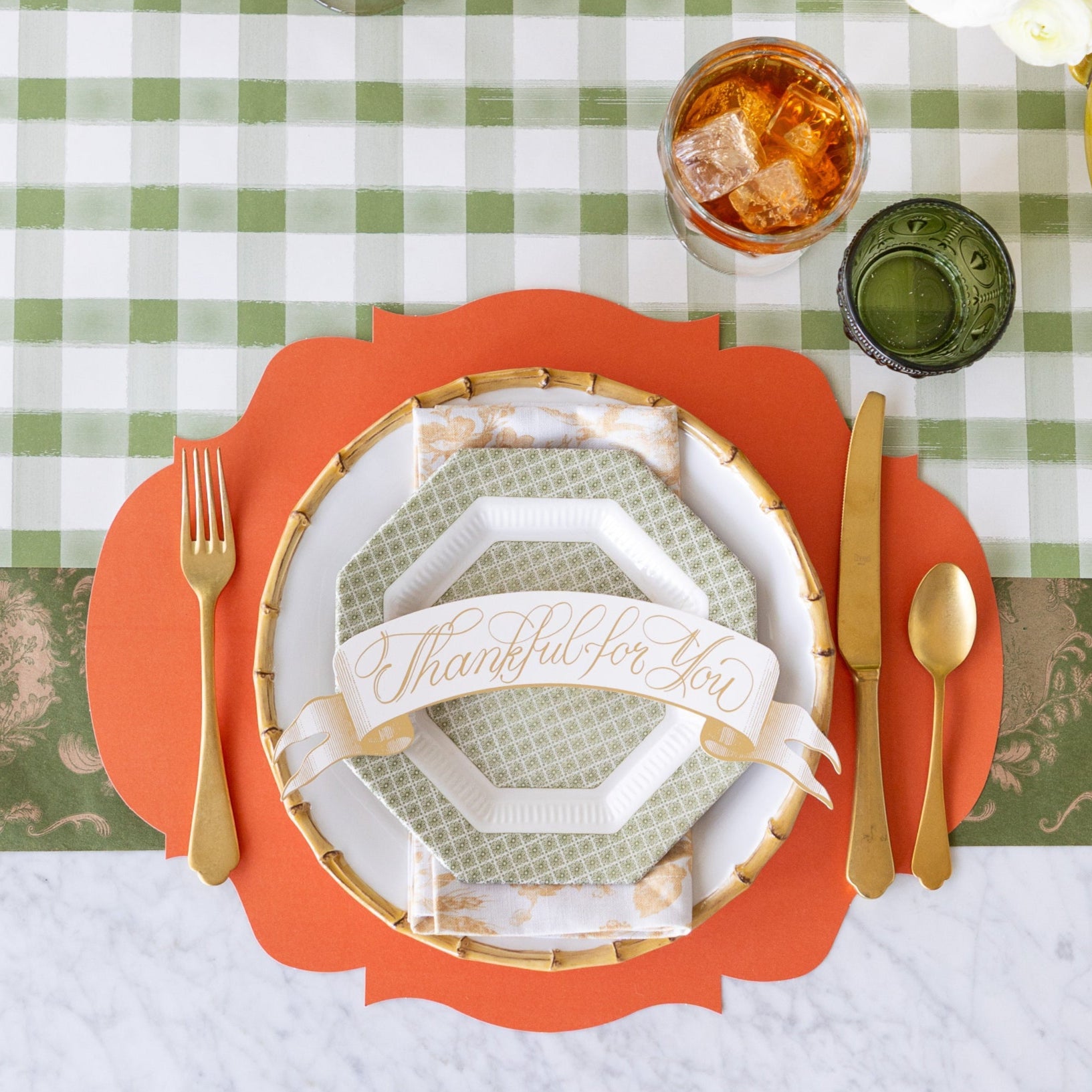 A fall table place setting with the Die-cut Orange French Frame Placemat, bamboo rimmed and vintage plates, Thankful For You Banner Table Accent, gold flatware, and iced tea on the Moss Painted Check and Moss Fable Toile Runner.
