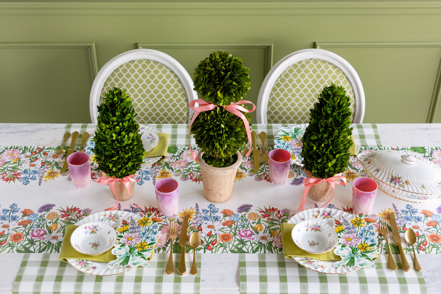 Dining table set with floral table runner, decorative topiaries, and place settings.