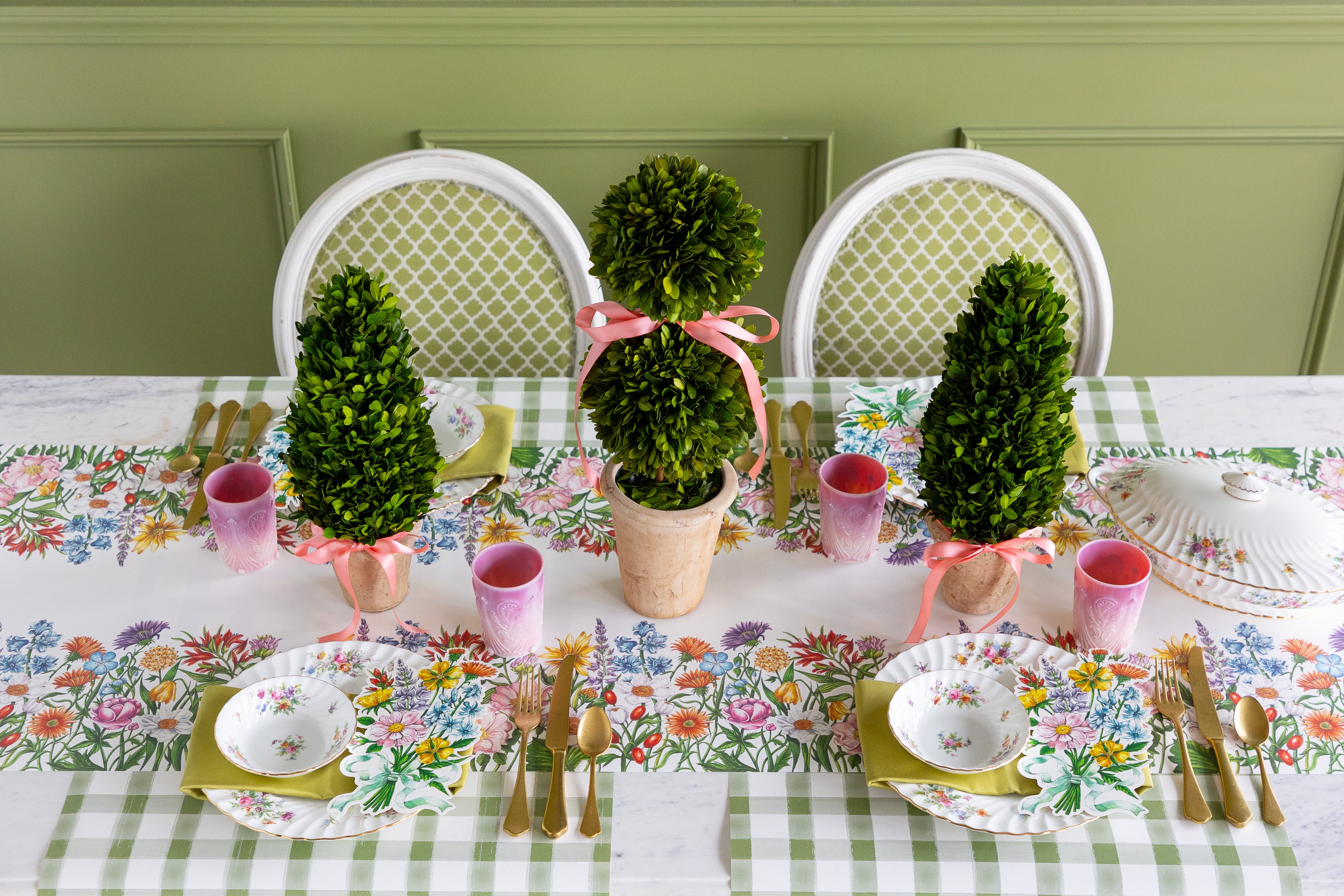 Dining table set with floral table runner, decorative topiaries, and place settings.