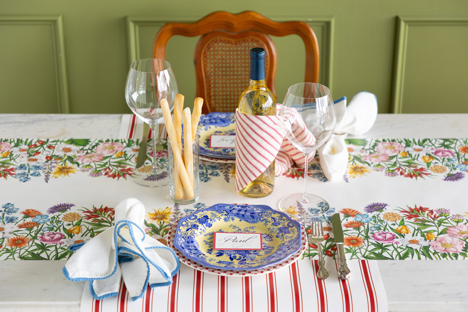 Set dining table with floral tablecloth, plates, cutlery, and wine bottle.
