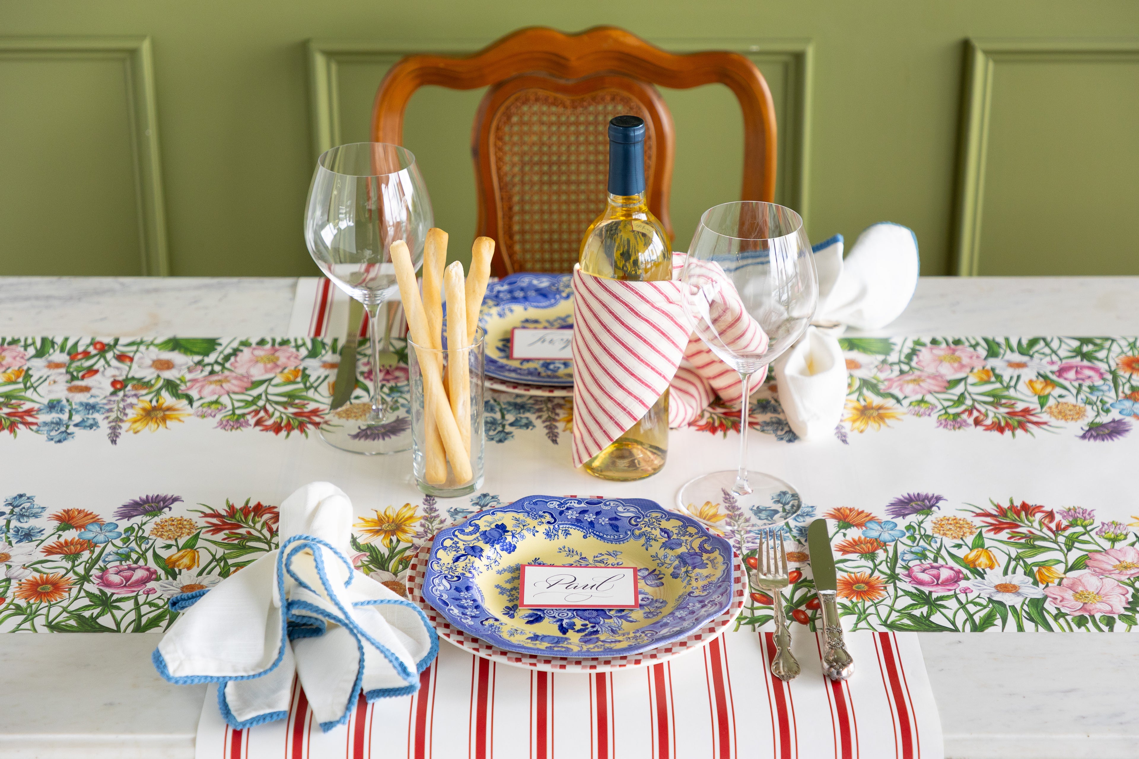 Set dining table with floral tablecloth, plates, cutlery, and wine bottle.