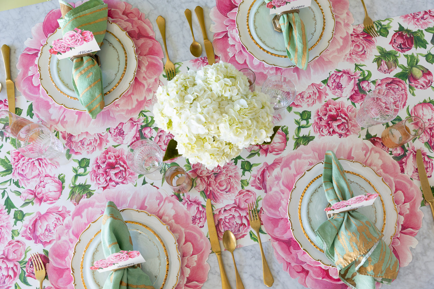 A decorative table setting with the Peonies in Bloom Runner, Die-cut Peony Placemats, decorative plates, Peonies Place Cards, marbled napkins and gold flatware.